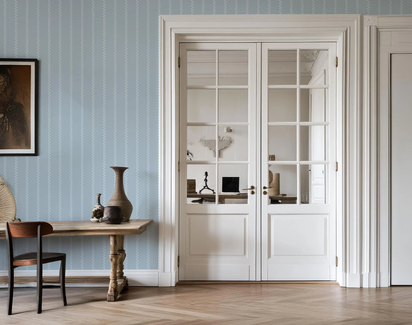 a room with a wooden table, a vase, and a chair. The room has a blue and white color scheme, with a large window and white doors.