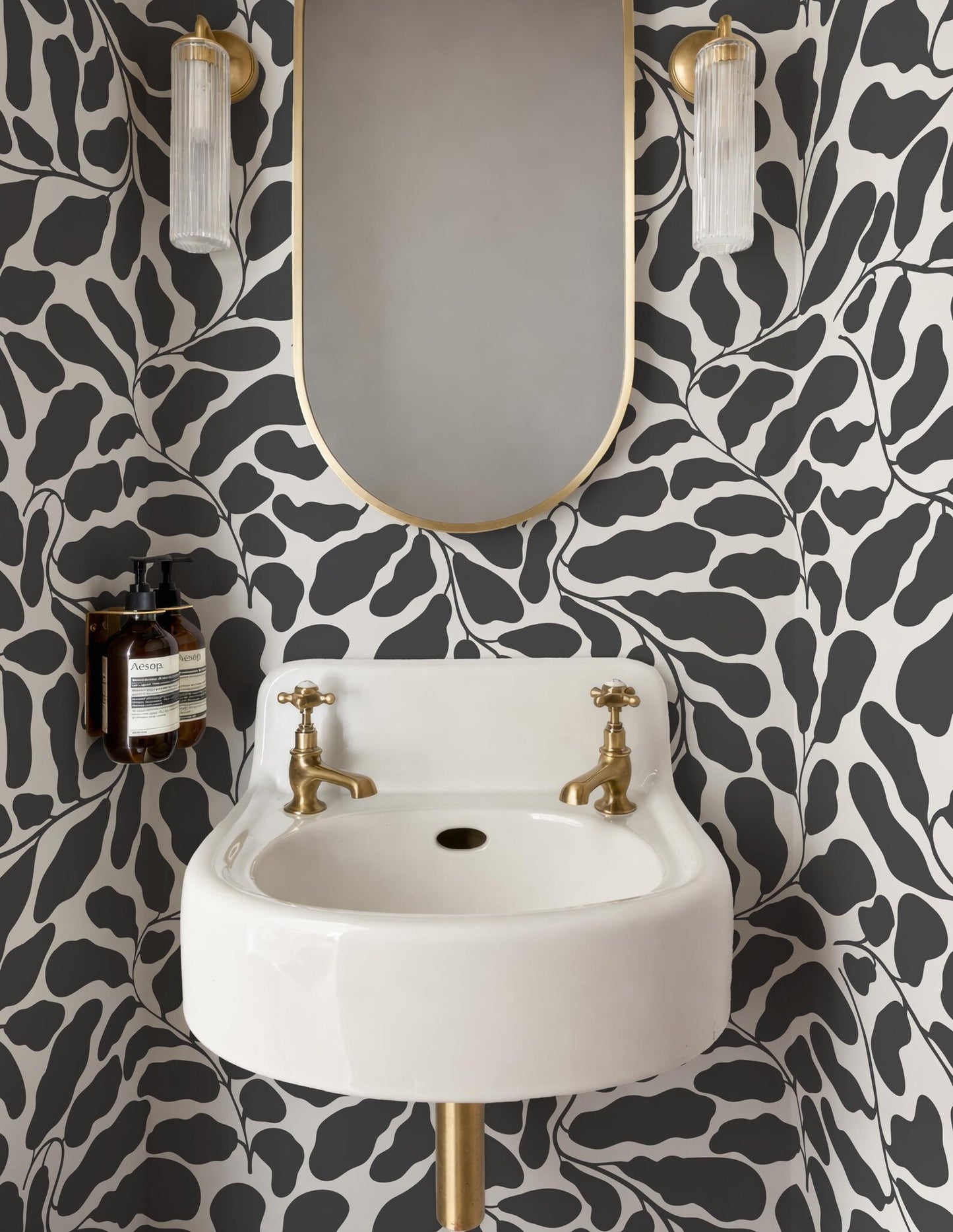 a bathroom with a white sink, a gold faucet, and a black and white leaf-patterned wall. Above the sink is a gold-framed mirror, and two light fixtures are mounted on either side of the mirror.
