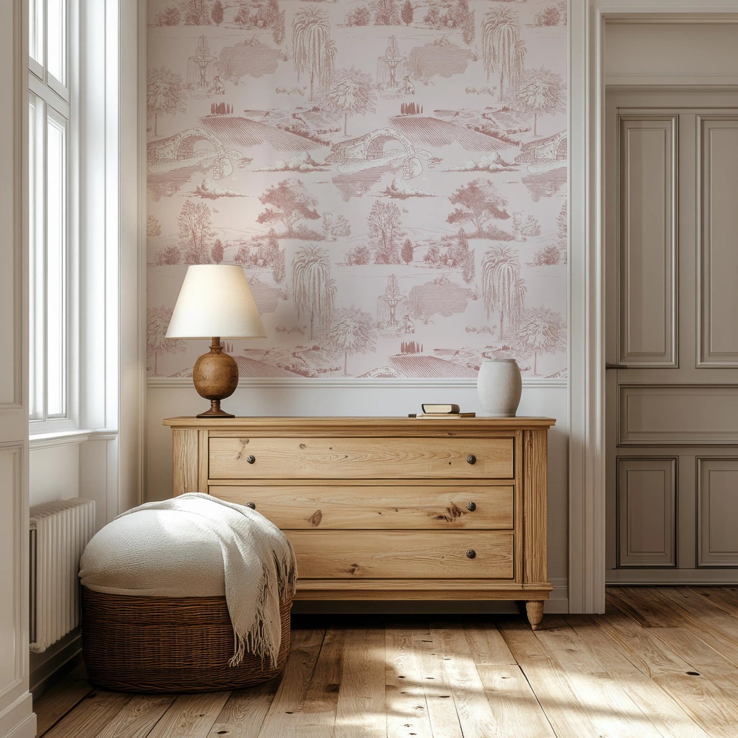 a cozy corner of a room with a wooden dresser, a lamp, and a woven ottoman. The walls are adorned with a pink and white wallpaper featuring a landscape scene.