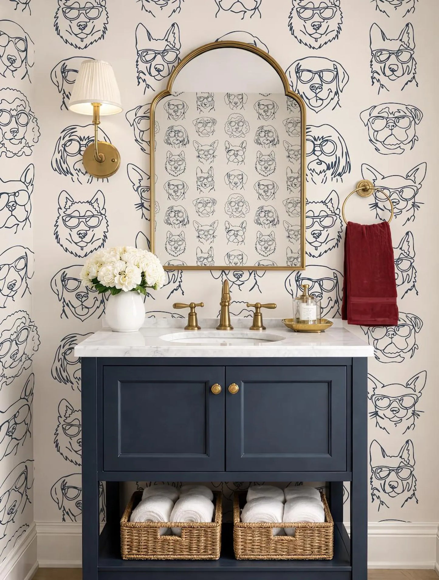 a bathroom with a blue vanity, a mirror with a gold frame, and a wallpapered wall with a repeating pattern of dog faces. The bathroom also includes a red towel, a white vase with flowers, and baskets for storage.