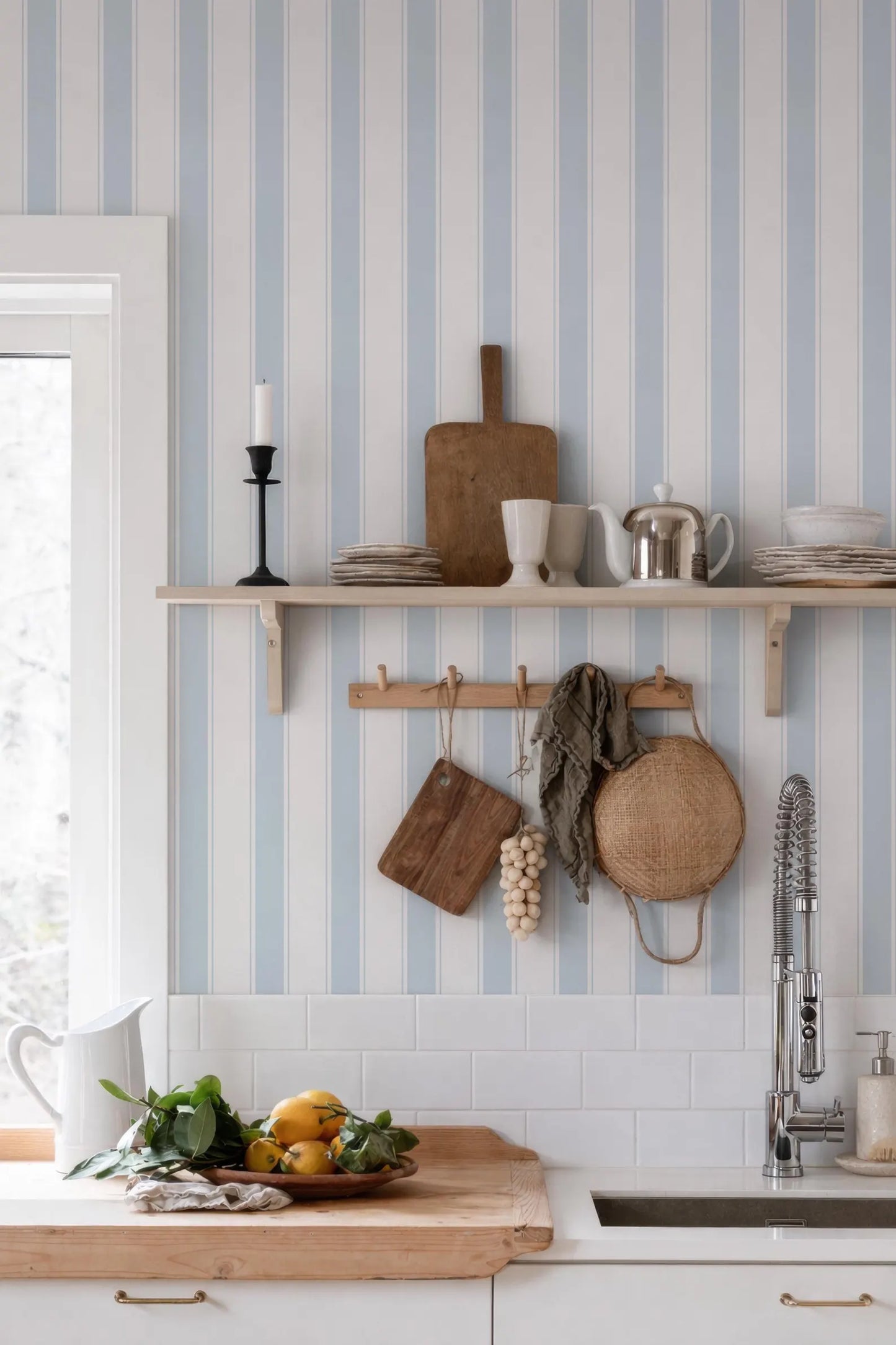 a kitchen with a wooden cutting board, a bowl of fruit, and a shelf with various kitchen items such as a teapot, a coffee pot, and a wooden spoon. The kitchen has a white countertop and a window, creating a bright and airy atmosphere.