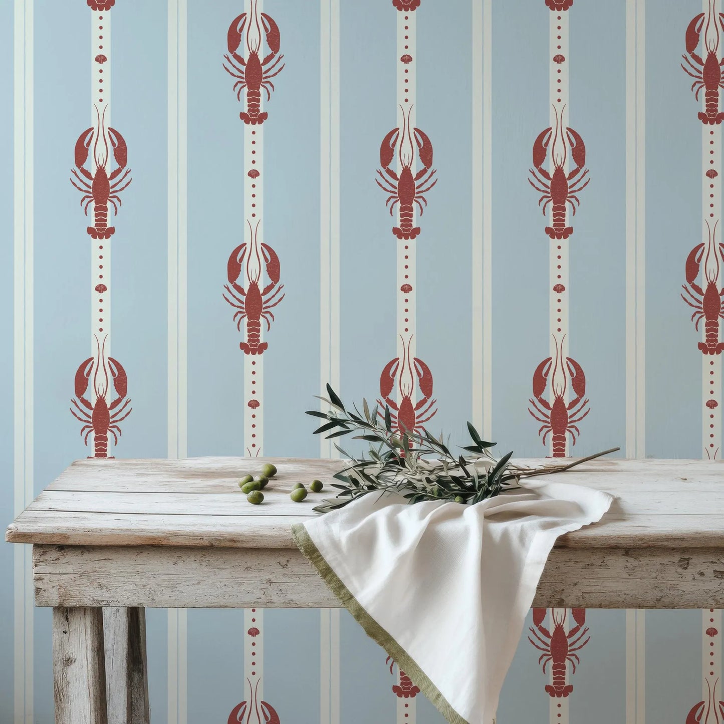 a white wooden table with a white cloth and a bunch of green olives, set against a blue and white striped wallpaper with red lobster designs.