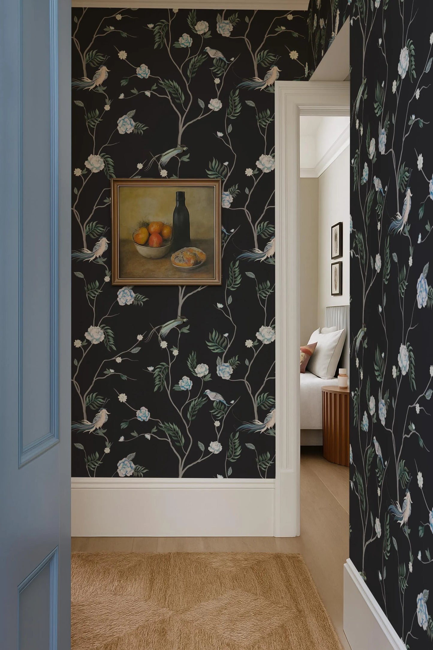 a hallway with a black and white floral wallpaper, a painting of a bowl of fruit on the wall, and a doorway leading to another room.