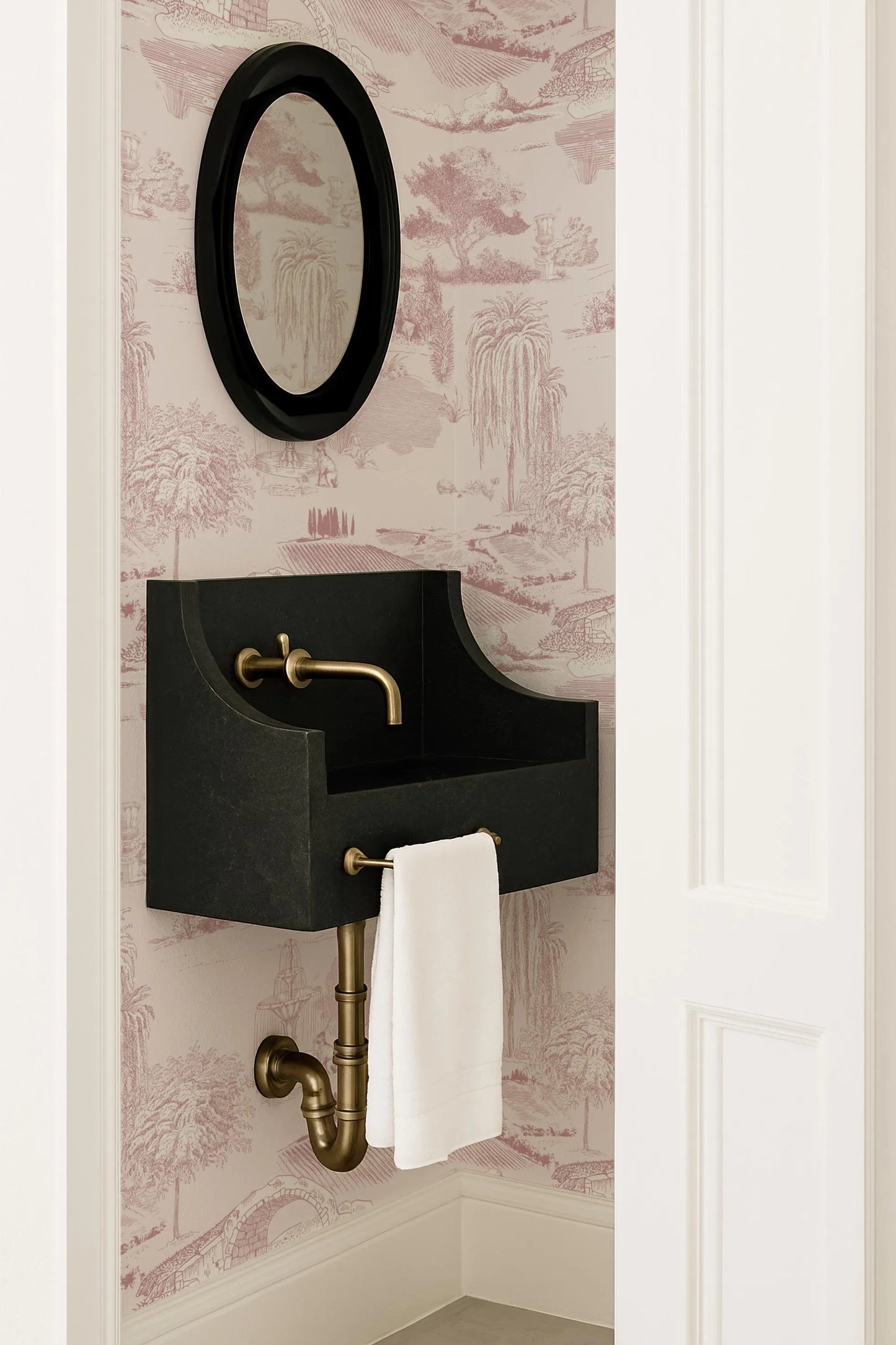 a bathroom with a black sink and a gold faucet, set against a pink and white patterned wallpaper. Above the sink is a black oval mirror with a gold frame.
