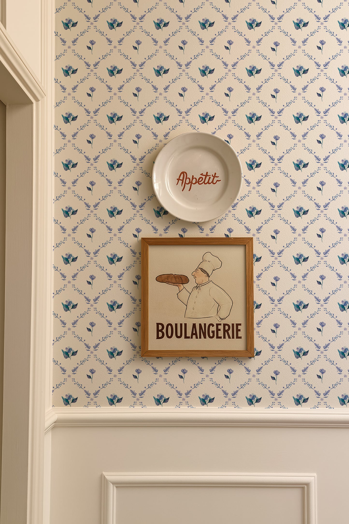 a wall with a framed picture of a chef holding a loaf of bread, a plate with the word "Appétit" written on it, and a wallpapered wall with a floral pattern.