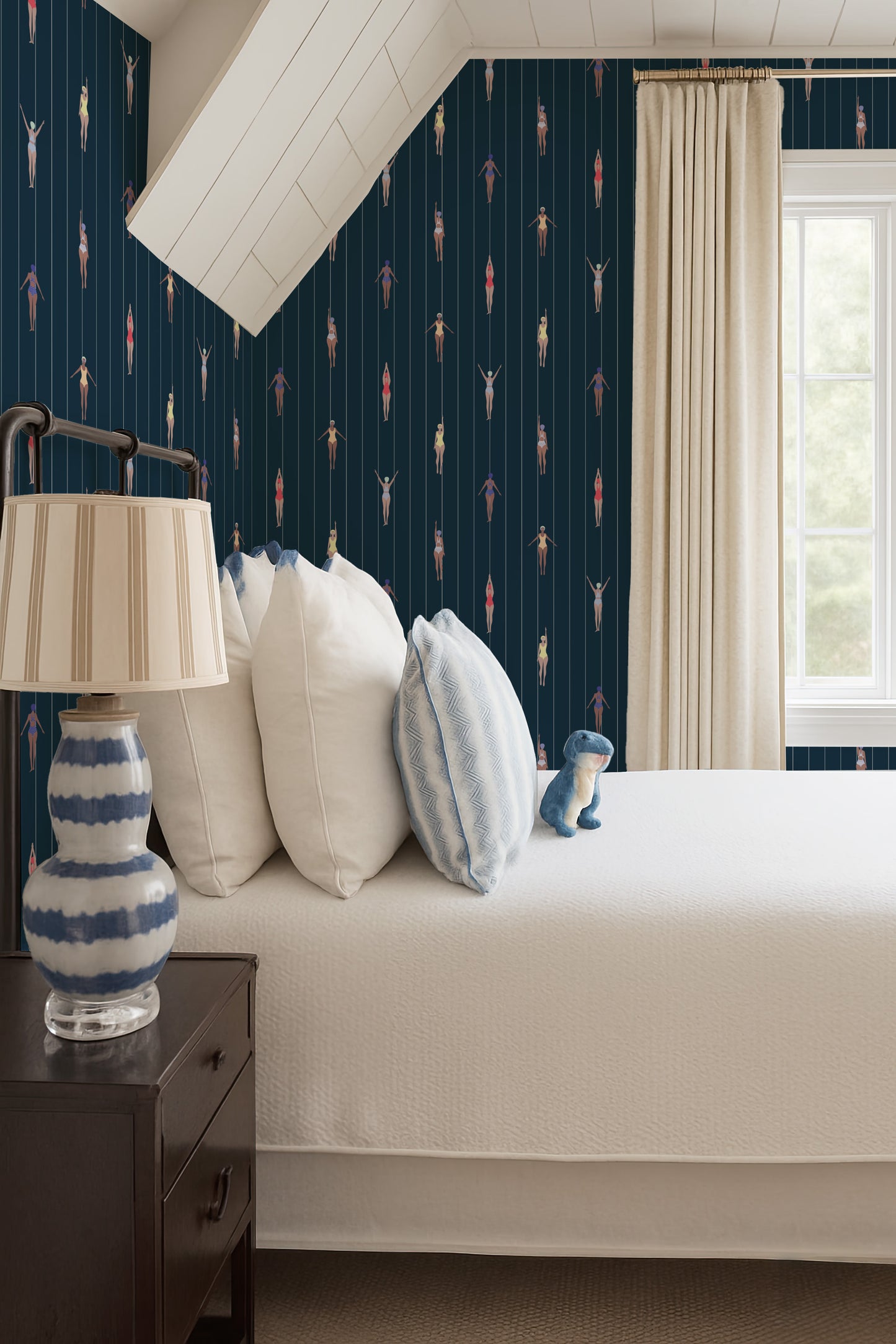 a bedroom with a bed, a nightstand, and a lamp. The bed has white pillows and a blue and white striped pillow. The nightstand has a lamp with a white shade. The wall behind the bed has a patterned wallpaper with a repeating design of fish and other aquatic creatures.