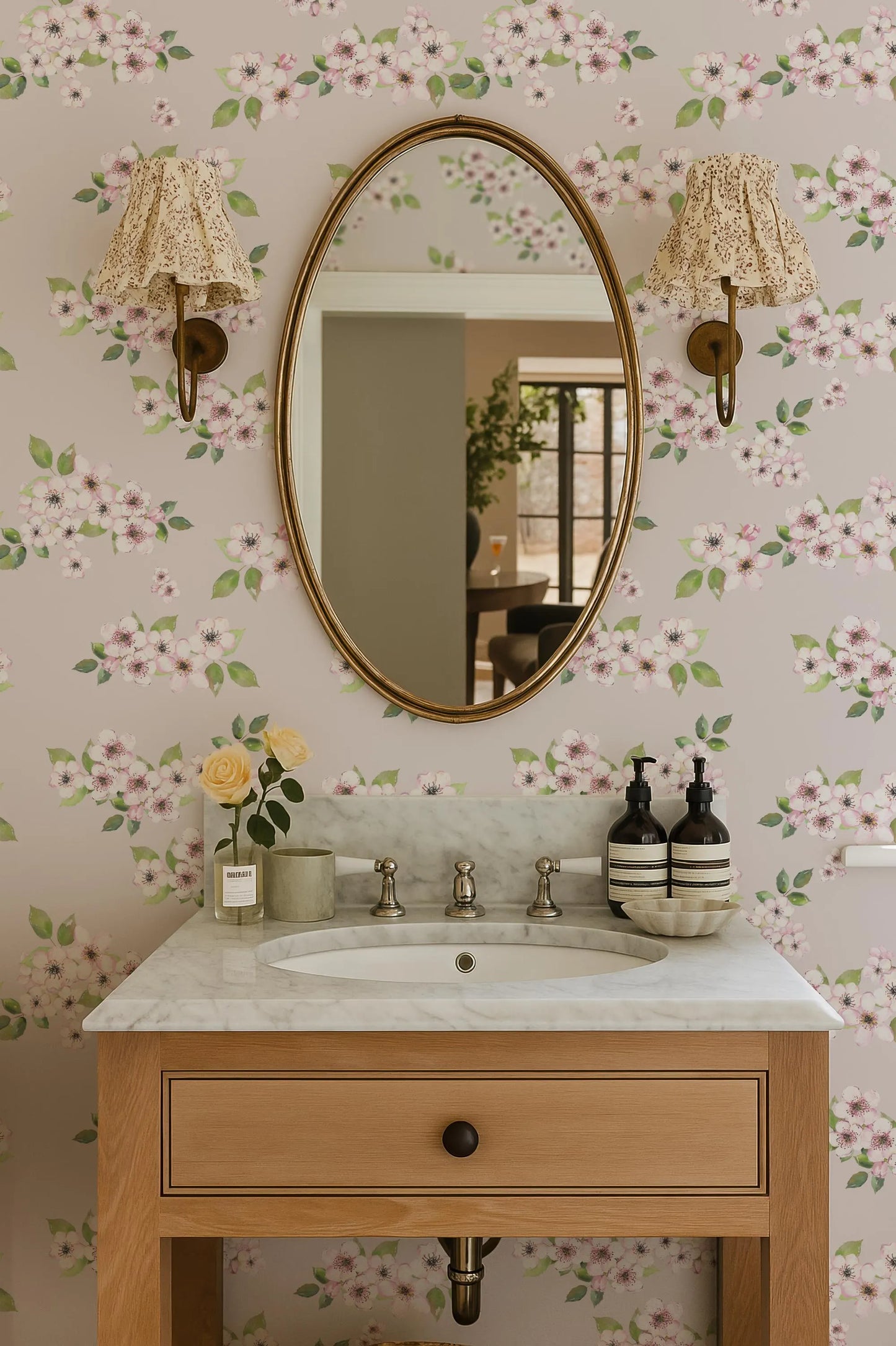 a bathroom sink with a marble countertop, a large oval mirror, and two wall sconces. The walls are decorated with a floral wallpaper pattern, and there are some decorative elements such as a vase of flowers and a bottle on the countertop.