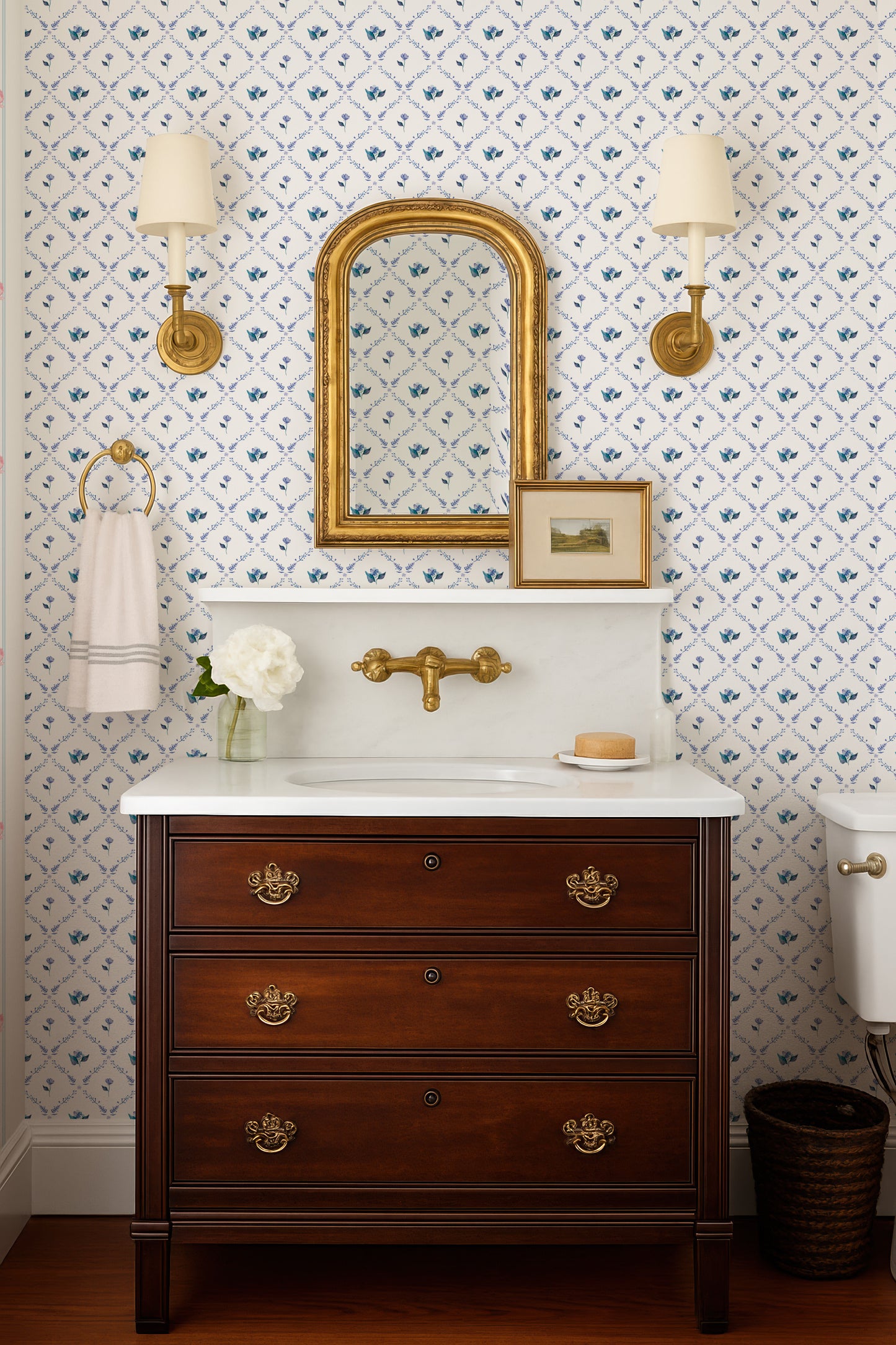 a bathroom with a wooden vanity, a white sink, and a mirror above it. The walls are decorated with blue and white floral wallpaper, and there are two wall sconces on either side of the mirror. The vanity has three drawers and a white countertop.