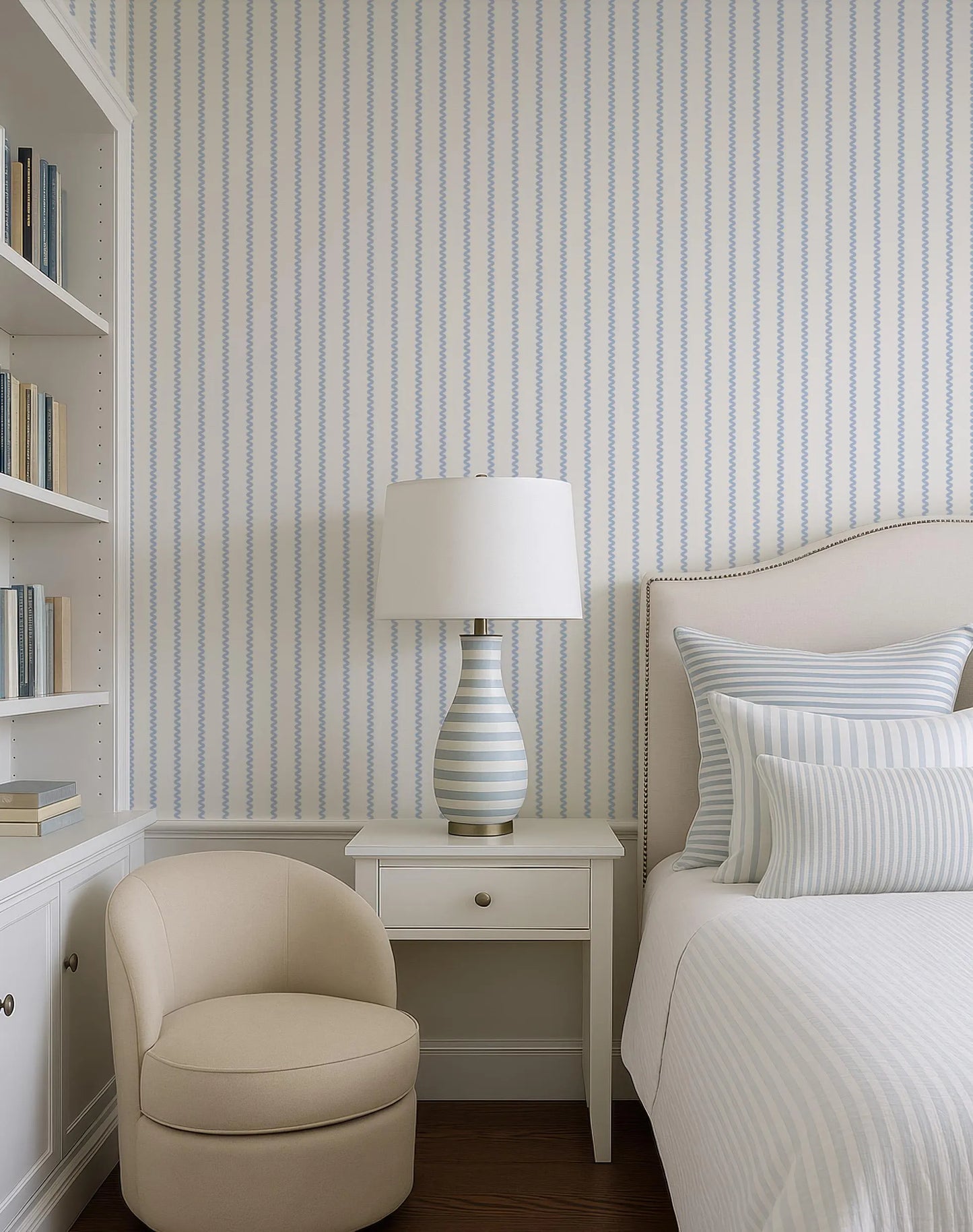 a cozy and well-organized bedroom with a bed, a white armchair, a white side table with a lamp, and a bookshelf filled with books.
