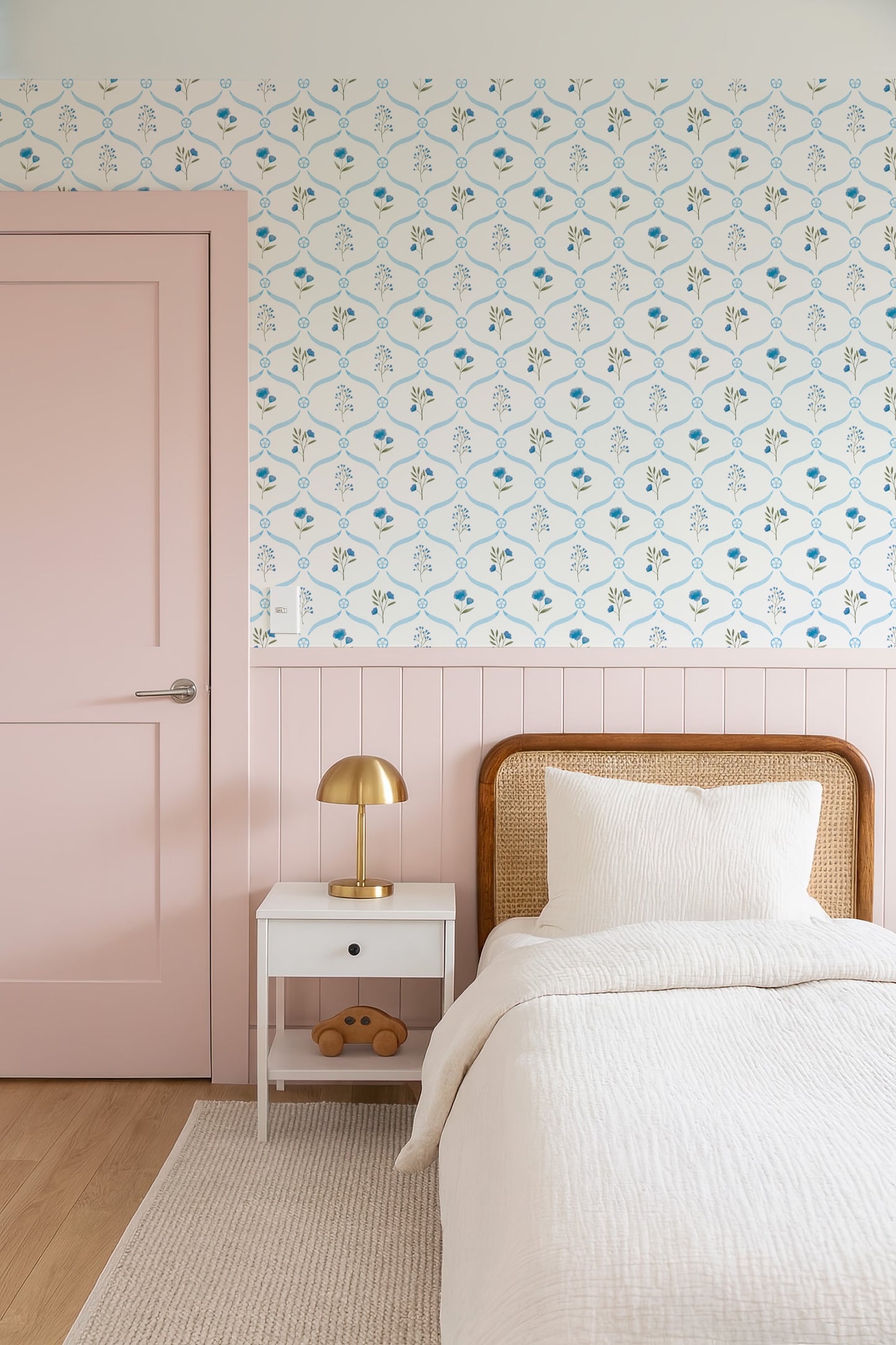 a cozy bedroom with a pink door, a white bed with a wooden headboard, and a white nightstand with a lamp. The walls are covered in a patterned wallpaper, and the overall color scheme is predominantly pink and white.