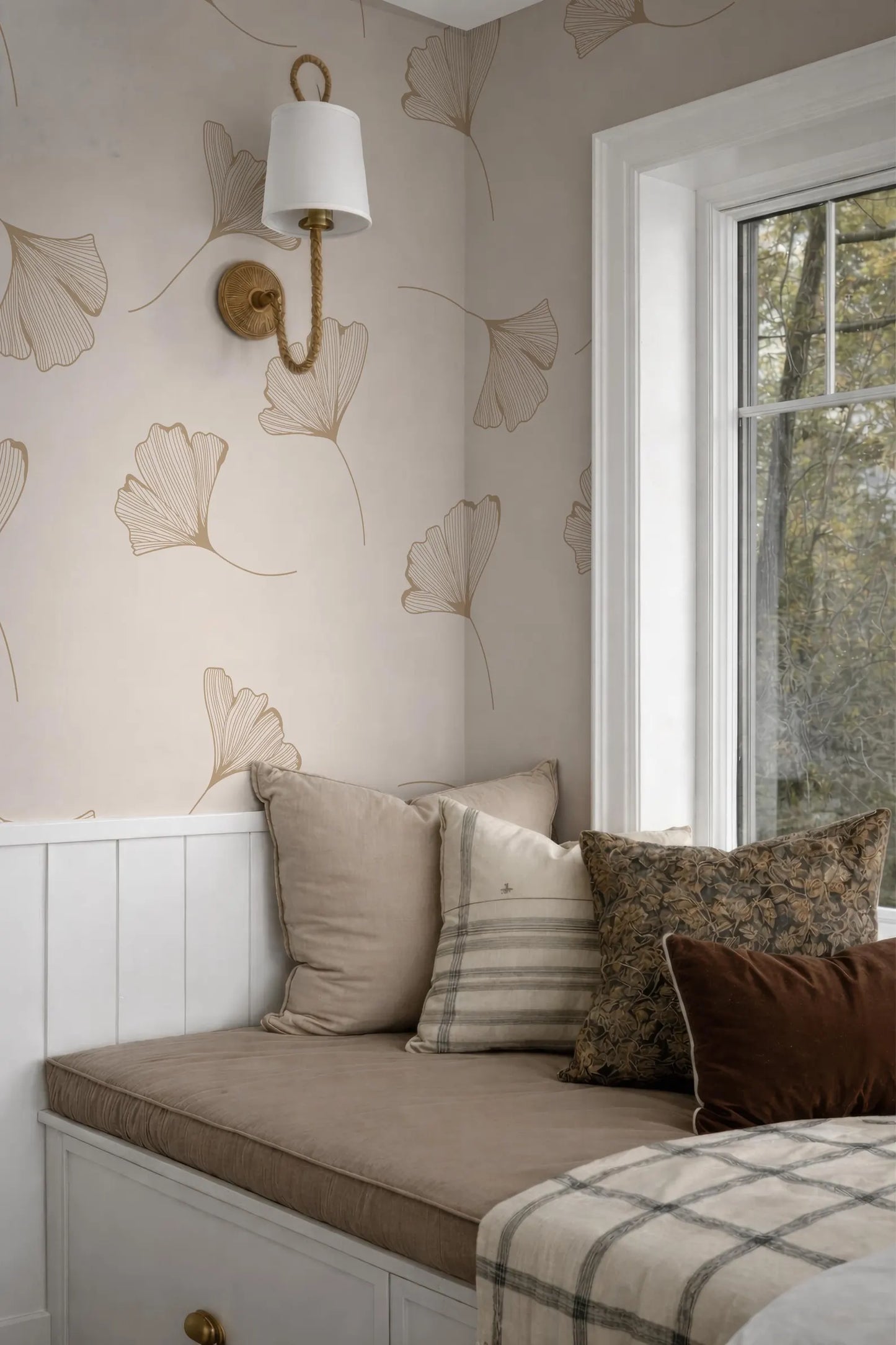 a cozy and inviting bedroom with a white wall adorned with a repeating pattern of ginkgo leaves. The room is furnished with a comfortable bed, a bench, and a window that allows natural light to enter the space. The bed is decorated with a striped pillow and a brown throw pillow, adding a touch of warmth and color to the room. The overall design and arrangement of the furniture create a harmonious and visually appealing atmosphere.