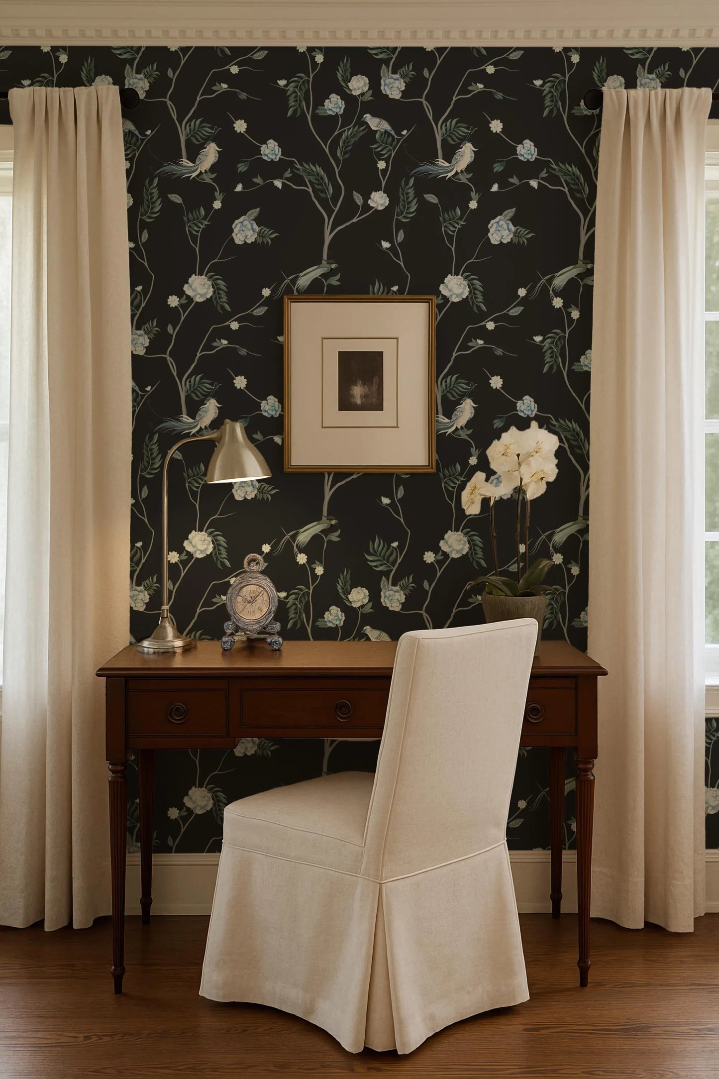 a cozy workspace with a wooden desk, a white upholstered chair, and a framed picture on the wall. The room has a dark floral wallpaper and a window with white curtains.