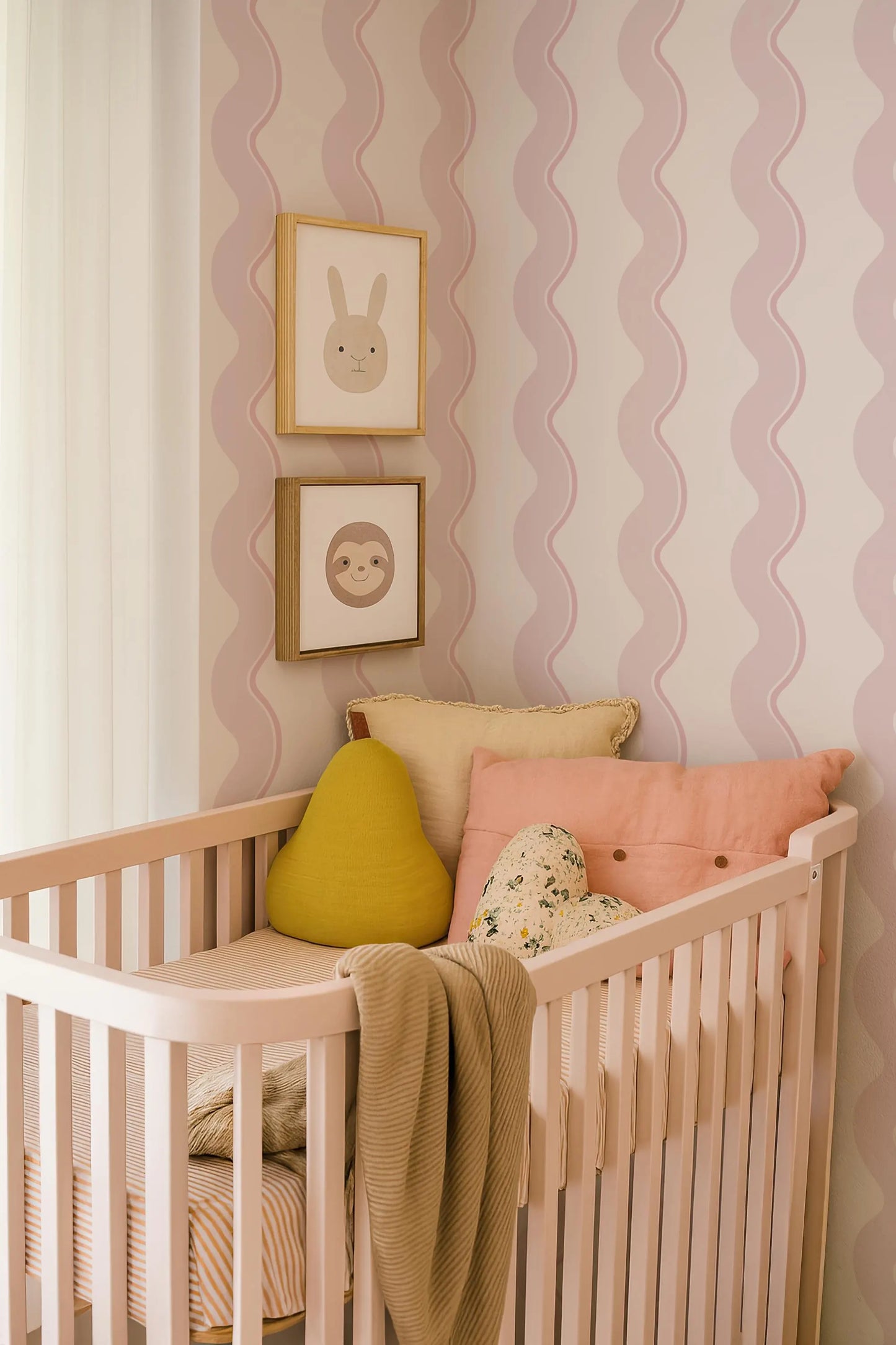 a cozy nursery with a white crib, adorned with a yellow pillow, a pink pillow, and a floral pillow. The crib is situated against a wall with pink and white striped wallpaper, and there are two framed pictures hanging above it.