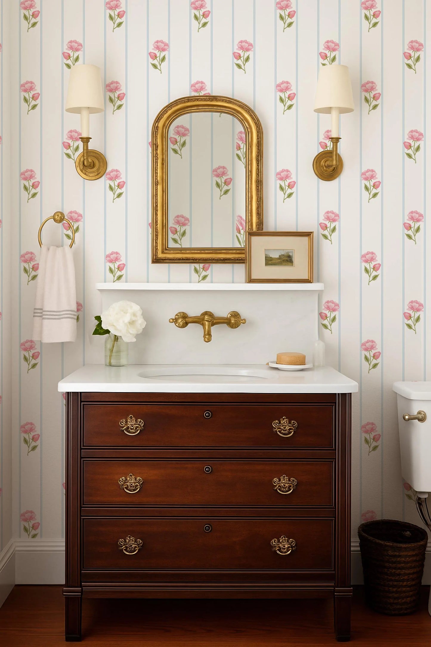 a bathroom with a wooden vanity, a white sink, and a mirror above it. The walls are decorated with pink and white floral wallpaper, and there are two wall sconces on either side of the mirror. The overall design has a classic and elegant aesthetic.