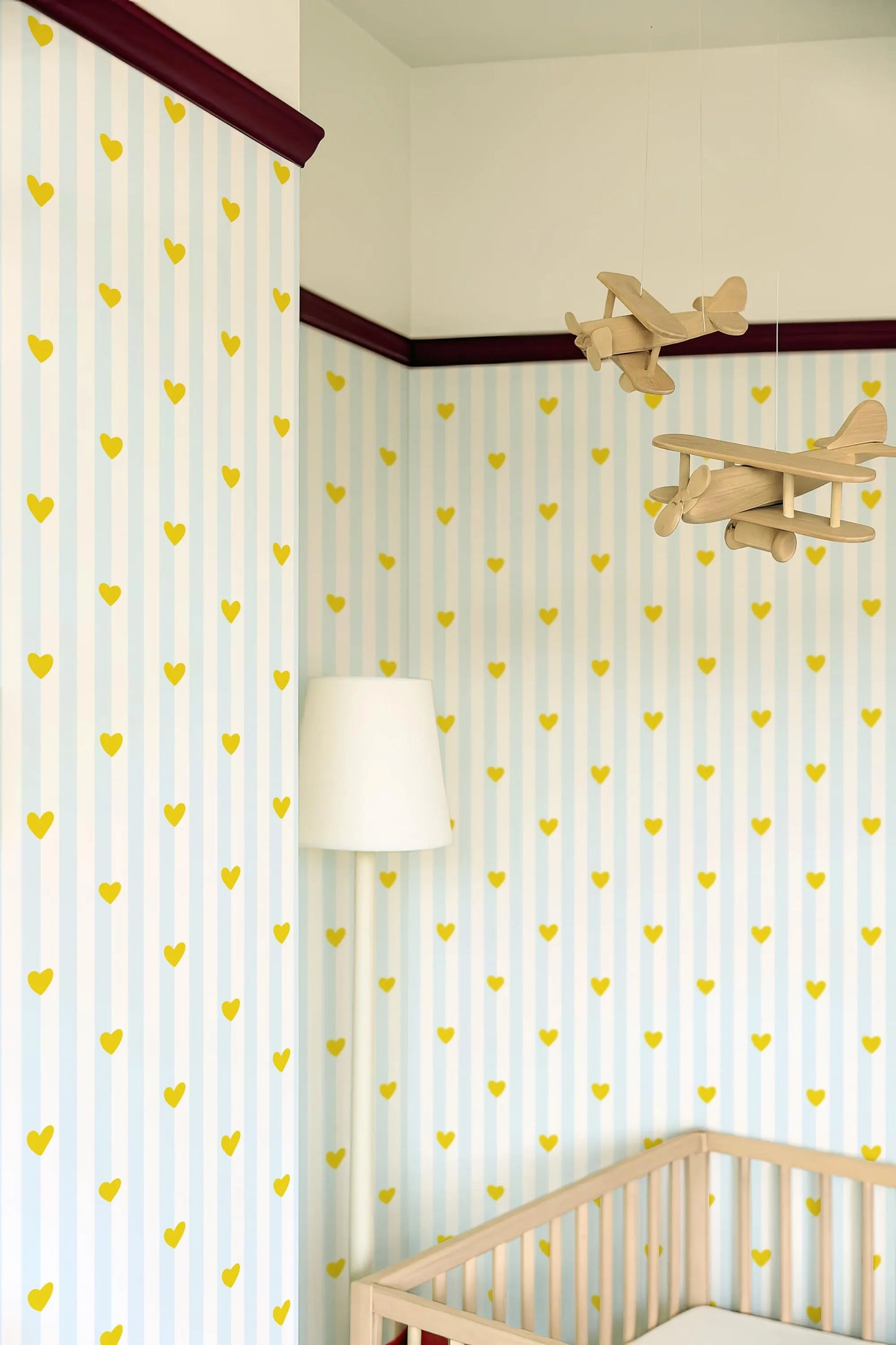 a nursery room with a crib, a white lamp, and a wooden airplane hanging from the ceiling. The walls are covered in a striped pattern with yellow hearts, creating a cheerful and playful atmosphere.