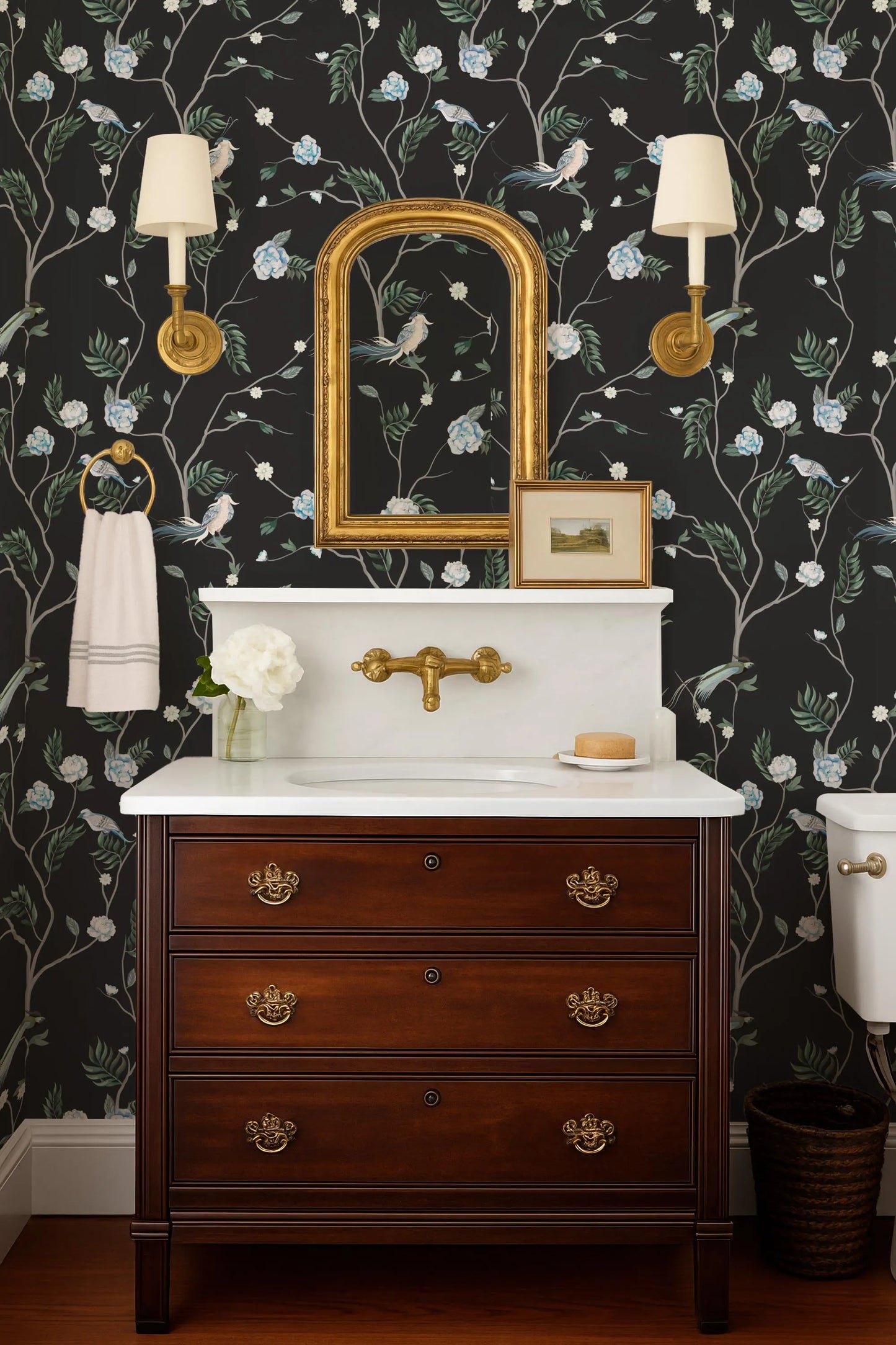a bathroom with a white sink, a wooden dresser, and a mirror above the sink. The walls are decorated with a floral wallpaper, and there are two wall sconces on either side of the mirror.