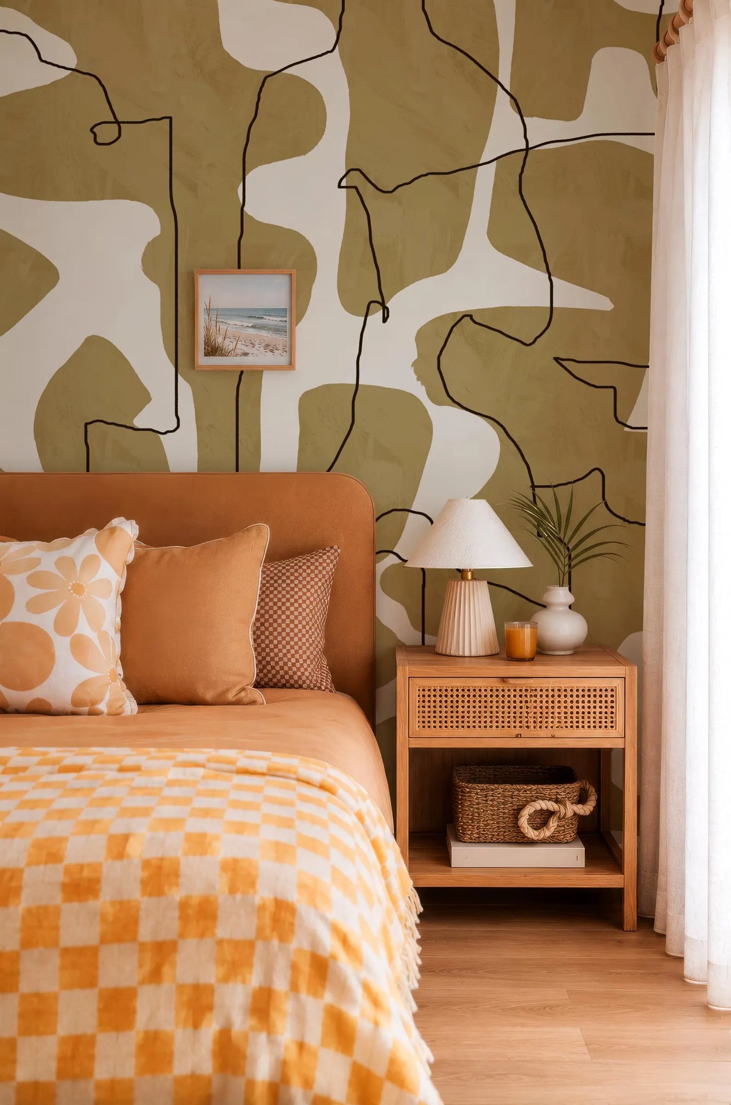 a cozy bedroom with a large bed adorned with a checkered bedspread and orange and white pillows. The bed is positioned against a wall with a bold abstract pattern in shades of green and white. A wooden nightstand with a lamp and a plant is situated next to the bed, and a basket is placed on the nightstand.