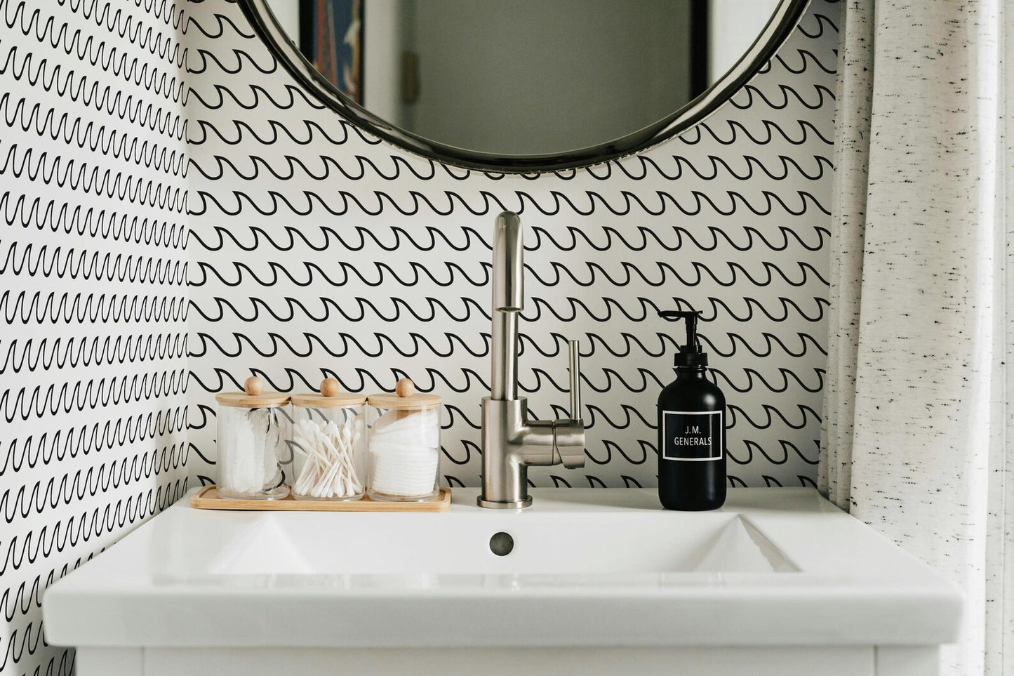 a bathroom sink with a black soap dispenser and a wooden tray holding cotton swabs and cotton buds. The sink is set against a wall with a black and white patterned wallpaper.