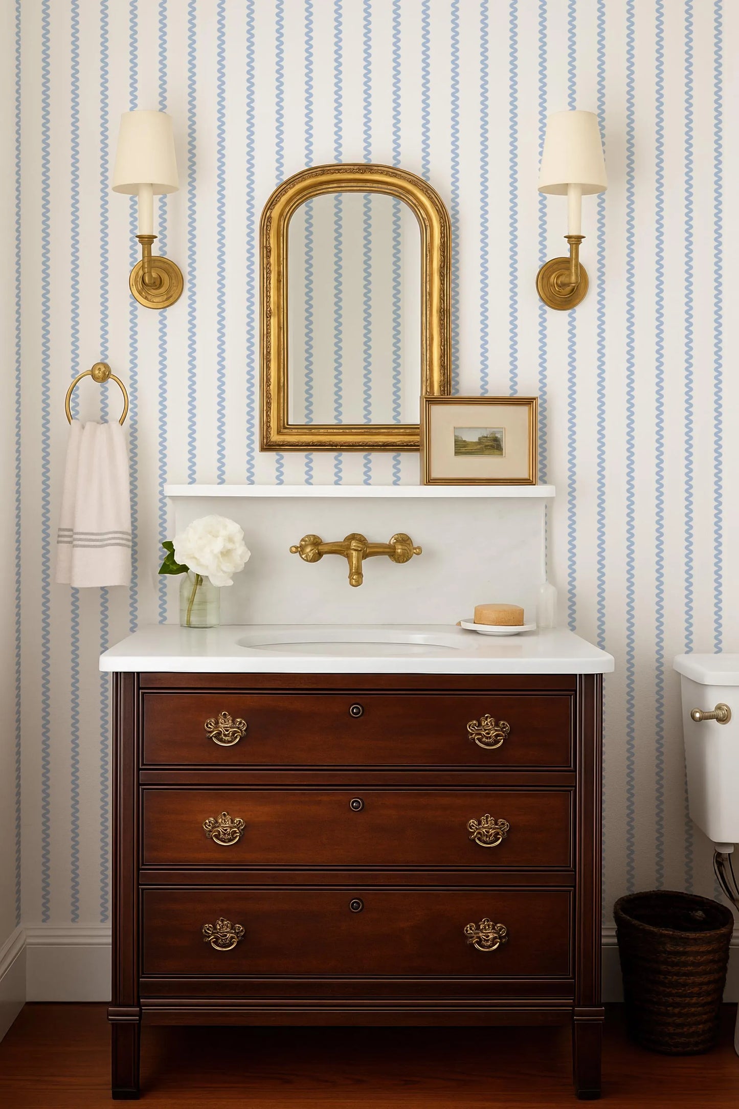 a bathroom with a wooden dresser with a sink and mirror above it. The dresser has three drawers and a white countertop. The walls are covered in blue and white striped wallpaper, and there are two wall sconces on either side of the mirror. A white towel is hanging on a hook next to the dresser, and a small vase with a flower is placed on the countertop.