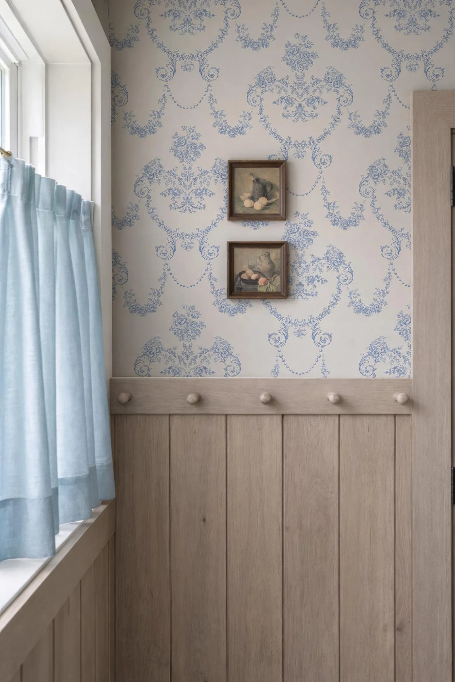 a cozy corner with a window, a wooden wall, and a wallpapered wall. There are two framed pictures hanging on the wall, and a curtain is partially drawn, allowing some light to enter the room.