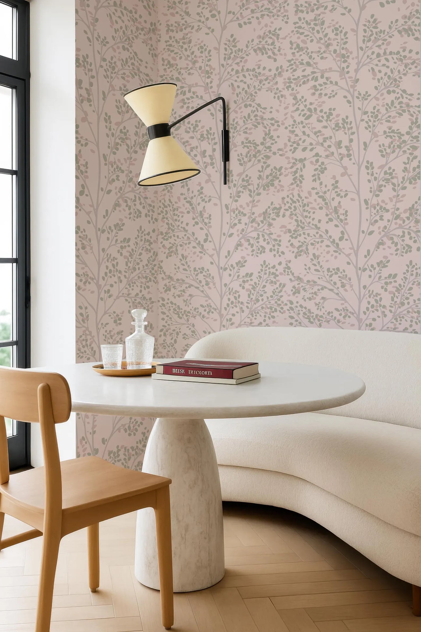 a modern and minimalist living space with a white sofa, a round white table, and a wooden chair. The walls are adorned with a pink and white floral wallpaper, and a lamp is hanging above the table. On the table, there is a book and a glass bottle.