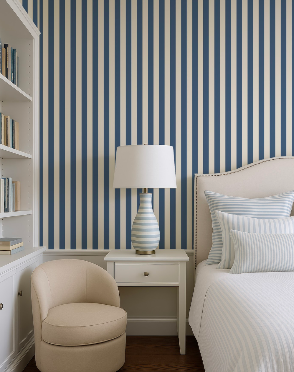a cozy bedroom with a white bed, a white armchair, and a white nightstand with a lamp. The room has a striped blue and white wallpaper, creating a calming and inviting atmosphere.