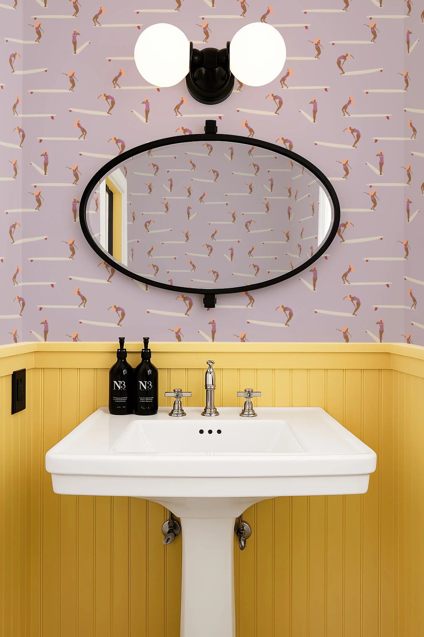 a bathroom with a white pedestal sink, a mirror above it, and a yellow wall with a patterned wallpaper. There are two bottles of soap on the sink.