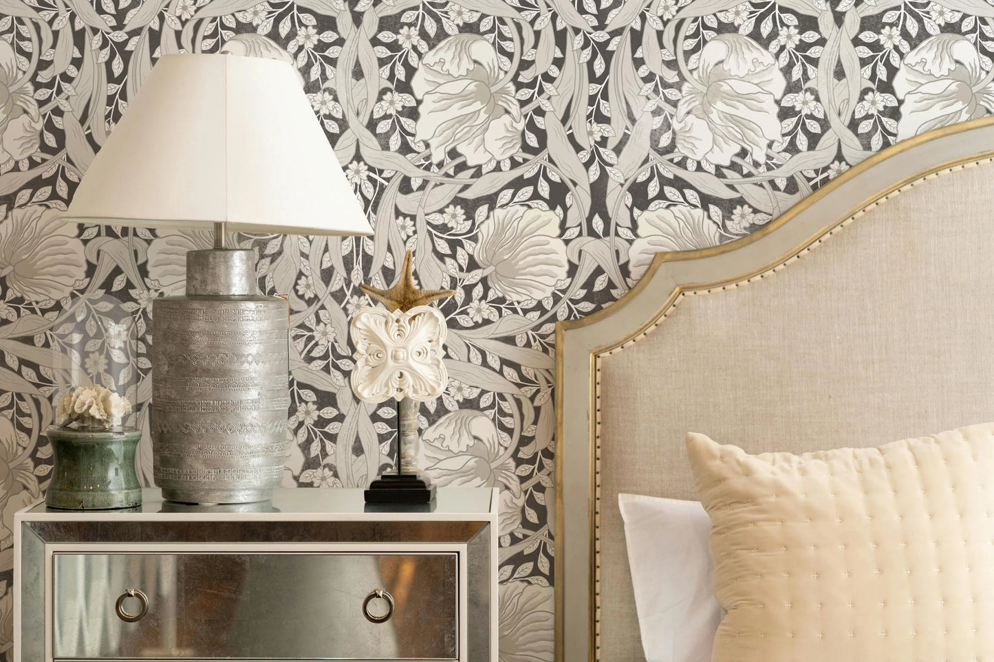 a bedroom setting with a bed, a dresser, and a lamp. The bed has a headboard and a white pillow, and the dresser has a mirror and a vase on top. The lamp is placed on the dresser, and the wall behind the bed has a floral wallpaper.