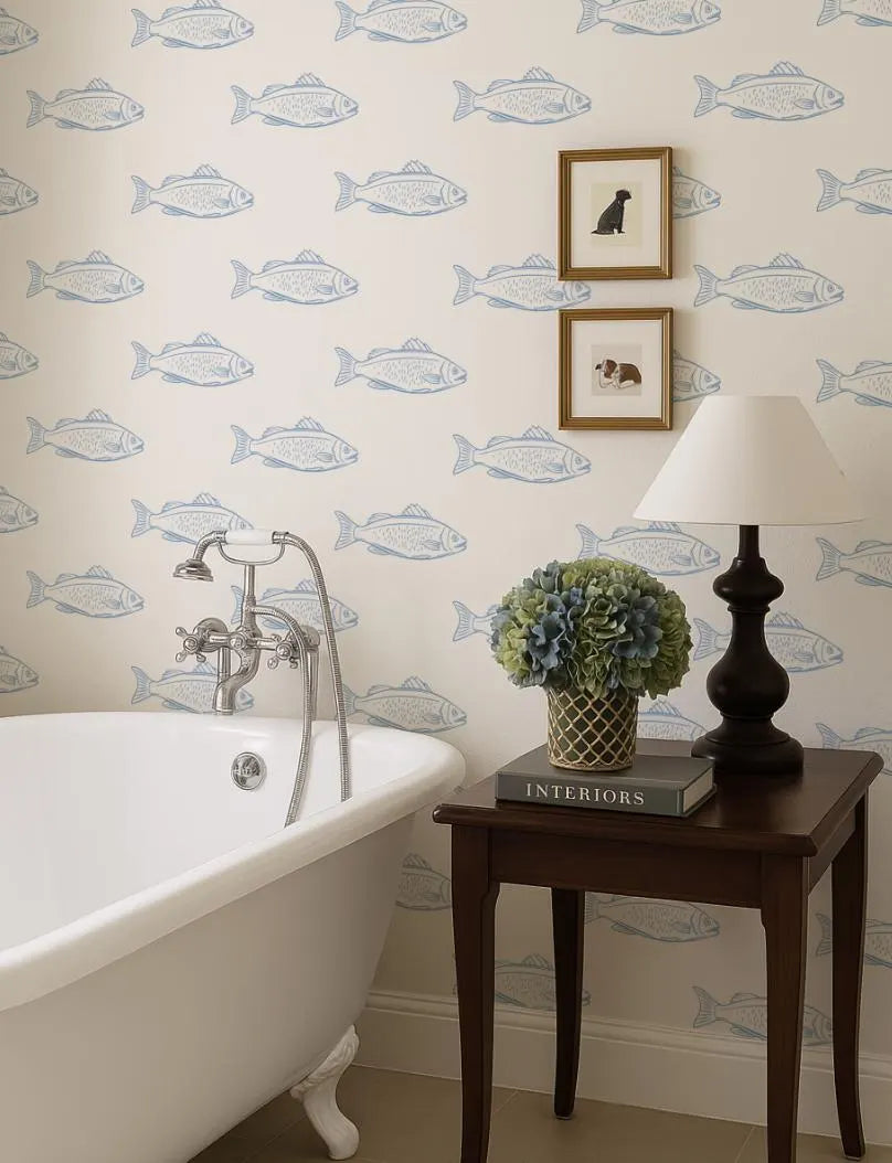 a bathroom with a white clawfoot bathtub, a wooden side table with a lamp, and a framed picture of a dog on the wall. The walls are decorated with a wallpaper pattern of fish.