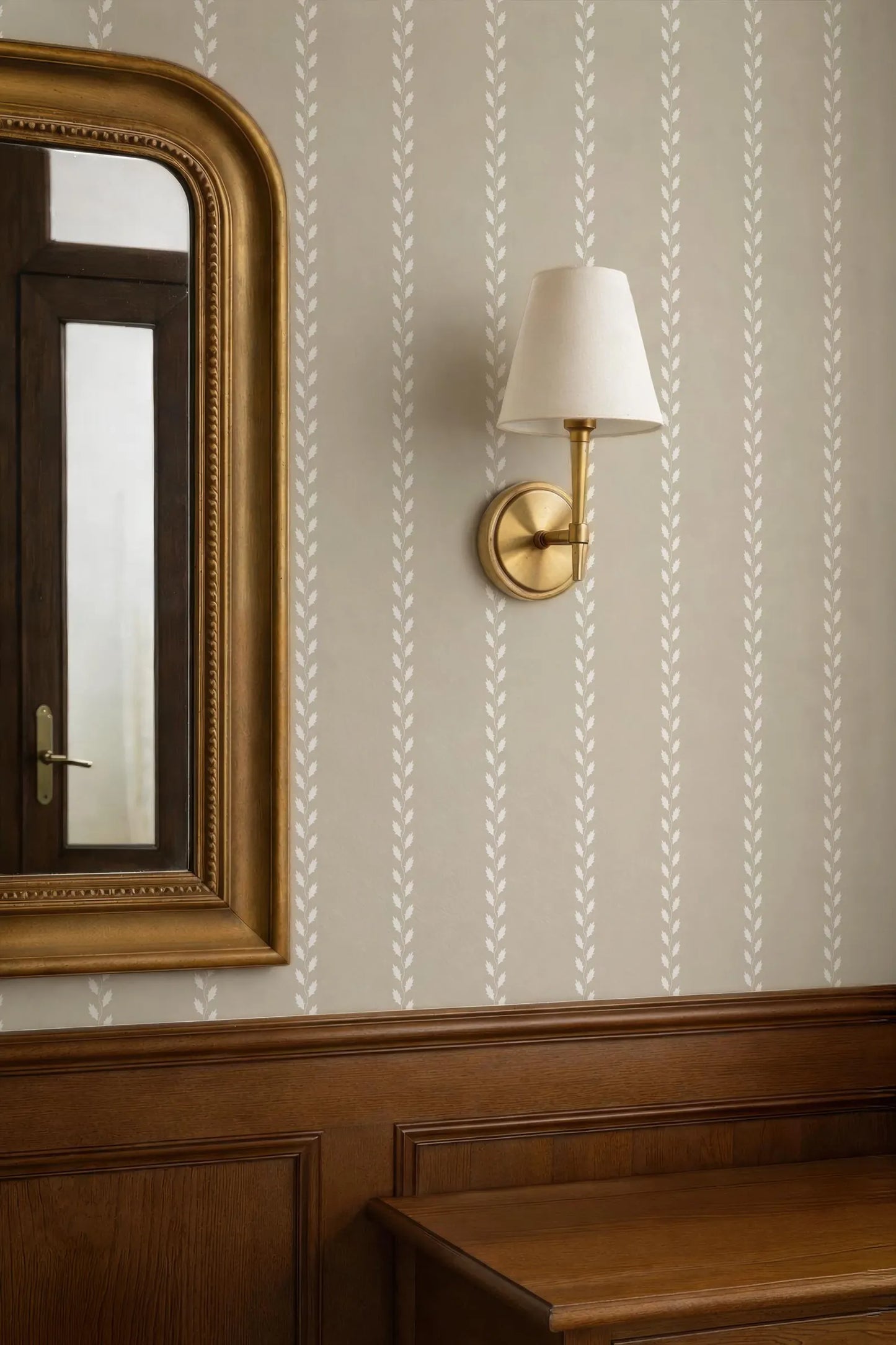 a wallpapered wall with a gold-framed mirror and a wall-mounted lamp with a white shade.