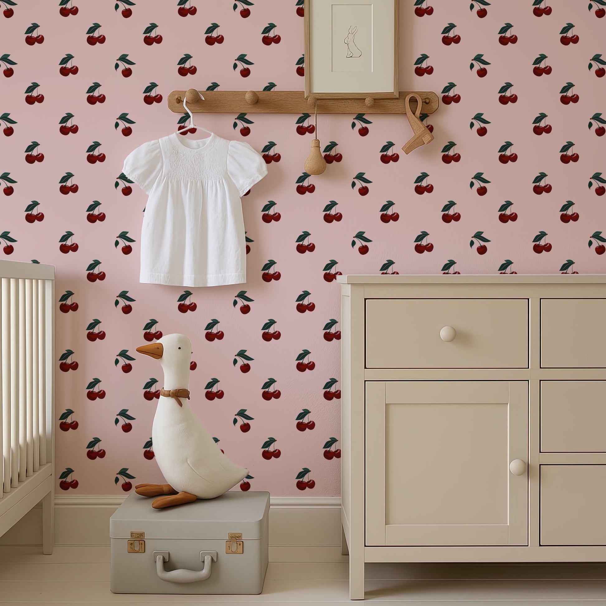 a nursery room with a pink wall decorated with a repeating pattern of cherries. A white dress is hanging on a wooden hanger, and a stuffed duck is sitting on a gray suitcase in the foreground.