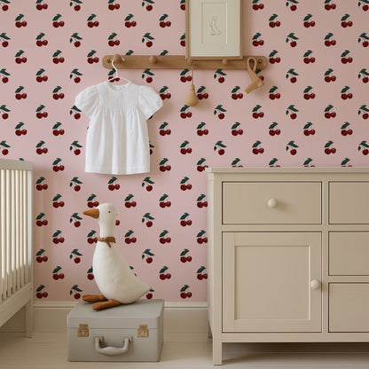 a nursery room with a pink wall decorated with a repeating pattern of cherries. A white dress is hanging on a wooden hanger, and a stuffed duck is sitting on a gray suitcase in the foreground.