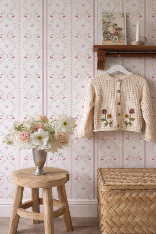 a cozy and charming interior with a wooden stool, a vase of flowers, a wooden shelf with a sweater, and a woven basket. The wall behind these items is adorned with a floral wallpaper pattern.