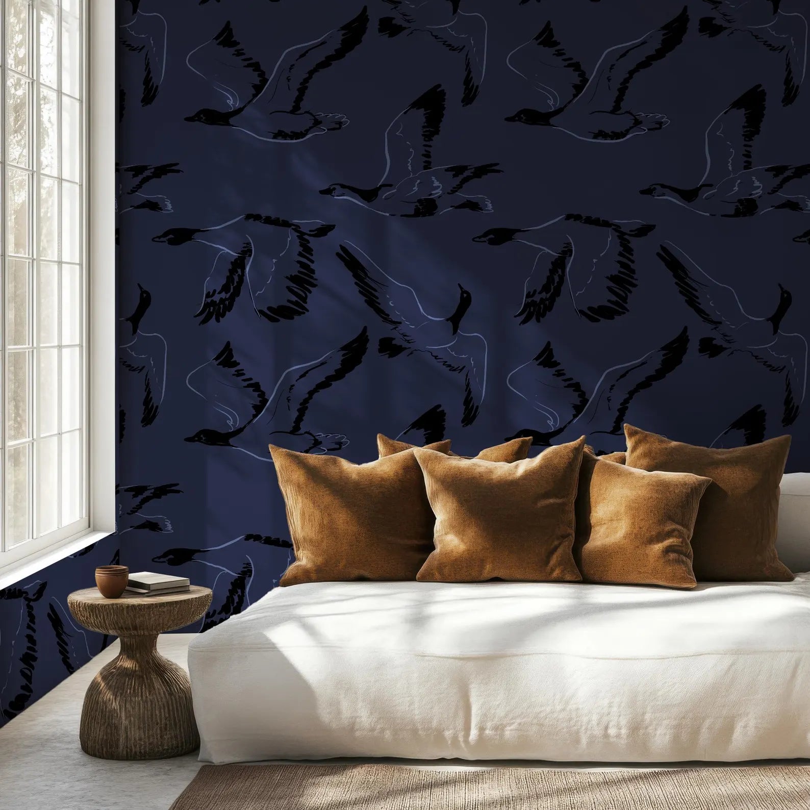 a cozy bedroom setting with a white bed adorned with brown throw pillows, a wooden side table, and a window with white curtains. The wall behind the bed is decorated with a dark blue pattern of black silhouettes of birds, creating a striking contrast against the white background.