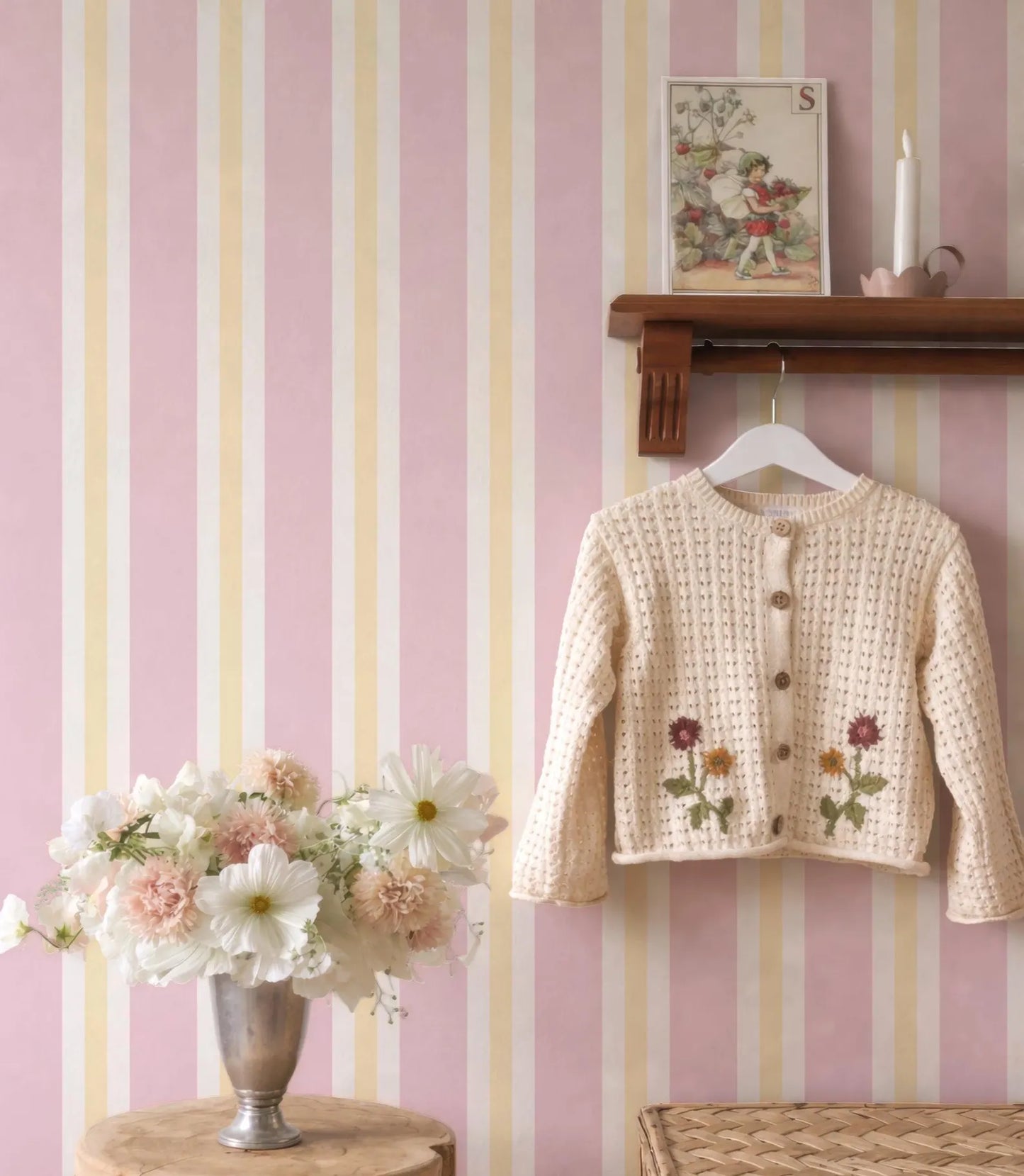 a cozy and charming scene with a pink and white striped wall, a wooden shelf displaying a sweater with flowers, and a vase of white and pink flowers.
