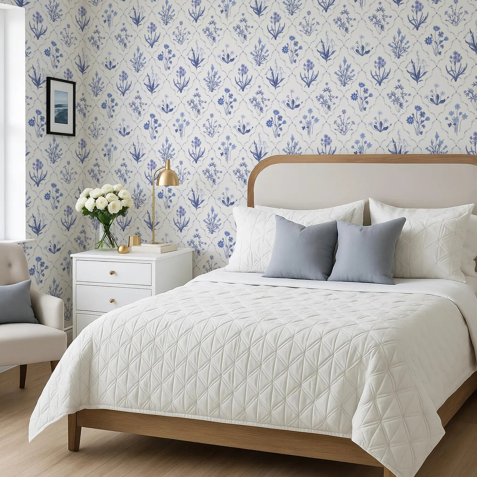 a bedroom with a large bed, a white headboard, and a blue and white floral wallpaper. The bed is covered in a white quilted comforter and has two pillows. There is a white nightstand next to the bed with a lamp and a vase of flowers. The room also features a chair and a window.
