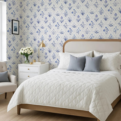 a bedroom with a large bed, a white headboard, and a blue and white floral wallpaper. The bed is covered in a white quilted comforter and has two pillows. There is a white nightstand next to the bed with a lamp and a vase of flowers. The room also features a chair and a window.