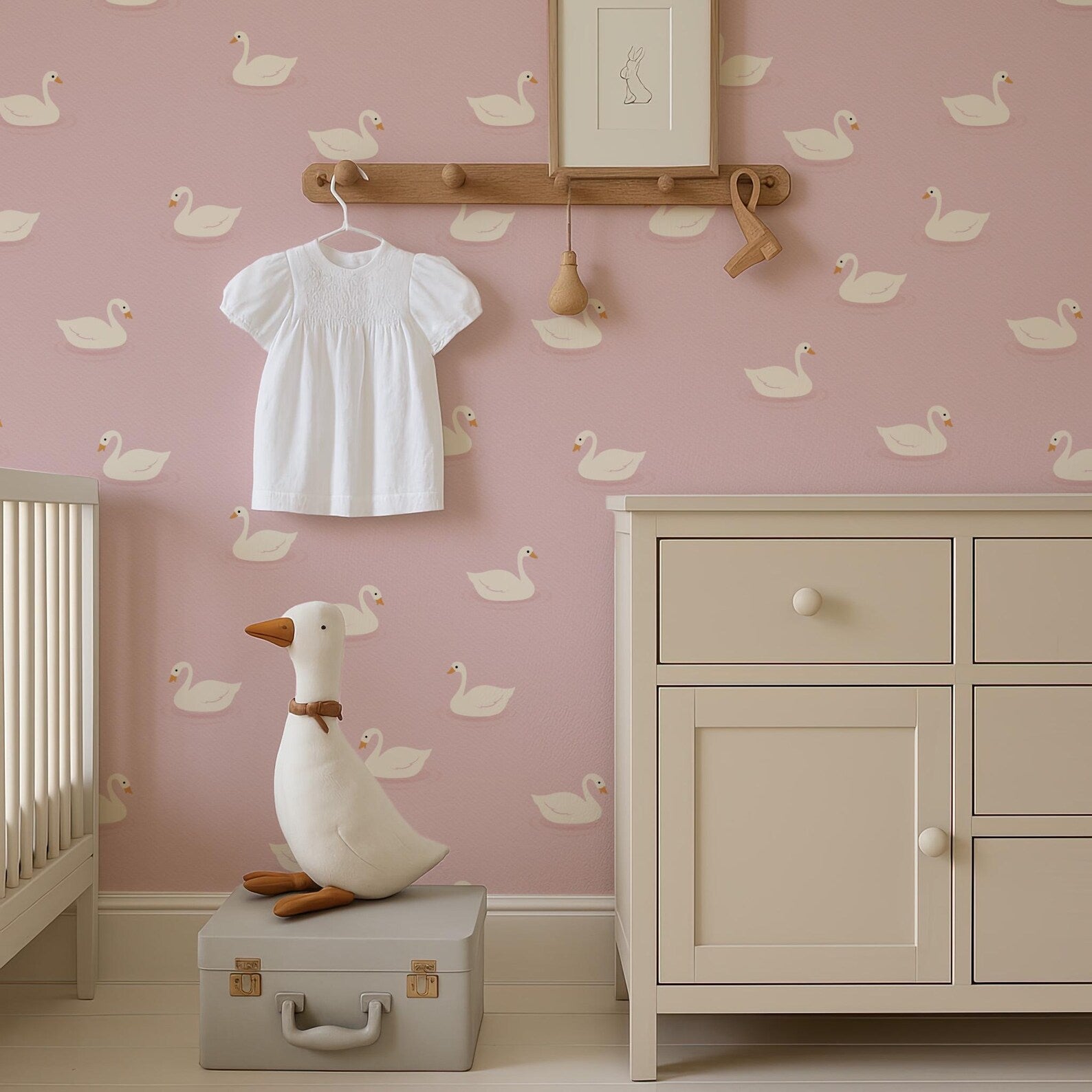 a nursery room with a pink wall decorated with white duck patterns. A white dress is hanging on a wooden hanger, and a white stuffed duck is sitting on a gray suitcase. A wooden shelf is mounted on the wall, and a white dresser is present in the room.