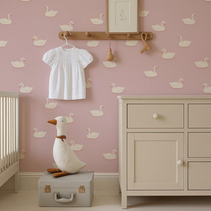 a nursery room with a pink wall decorated with white duck patterns. A white dress is hanging on a wooden hanger, and a white stuffed duck is sitting on a gray suitcase. A wooden shelf is mounted on the wall, and a white dresser is present in the room.