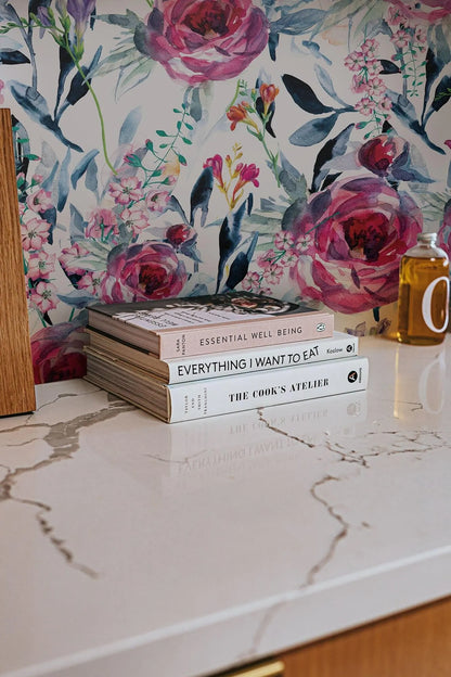 a white marble countertop with a stack of three cookbooks and a bottle of olive oil on it. The books are titled "Essential Well Being" and "The Cook's Aeti".