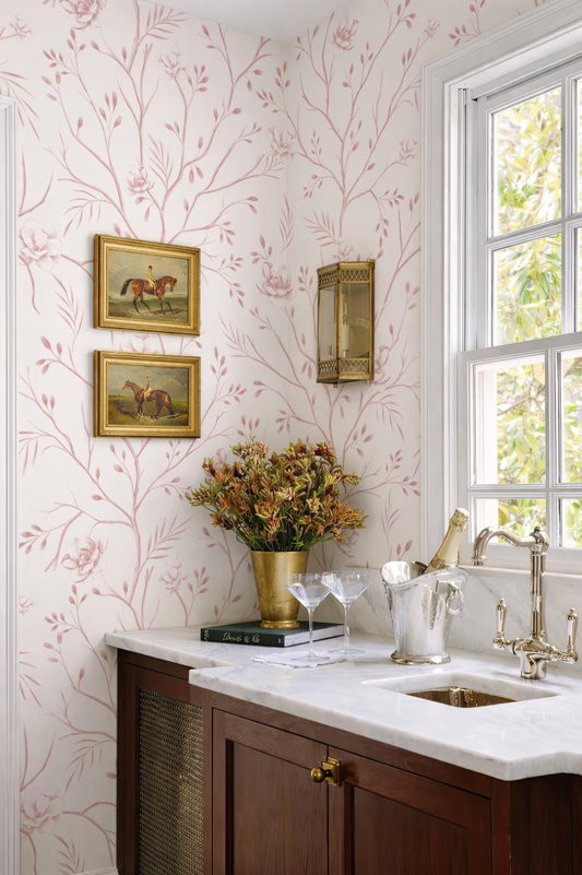 a cozy and elegant kitchen scene with a white countertop, a vase of flowers, and a wine glass. The walls are adorned with a pink floral wallpaper, and there are two framed pictures hanging above the counter. The kitchen also includes a sink and a window, allowing natural light to enter the space.