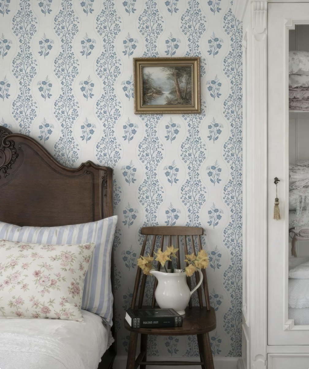 a cozy bedroom scene with a wooden headboard, a white and blue floral wallpaper, and a framed landscape painting on the wall. A wooden chair with a white pitcher of flowers and a book on it is also present in the room.