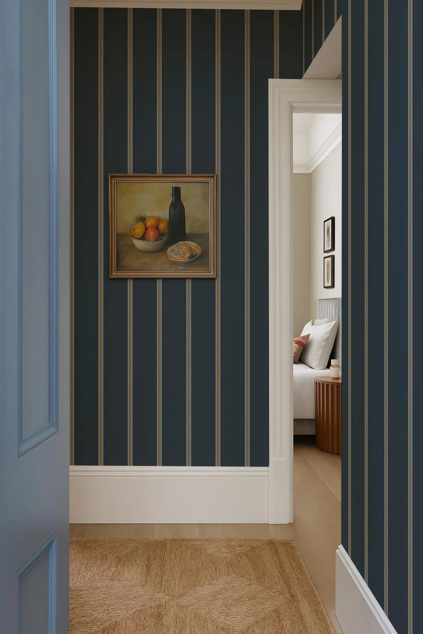 a hallway with a framed still life painting of fruit on the wall, a blue and white striped wallpaper, and a doorway leading to another room.