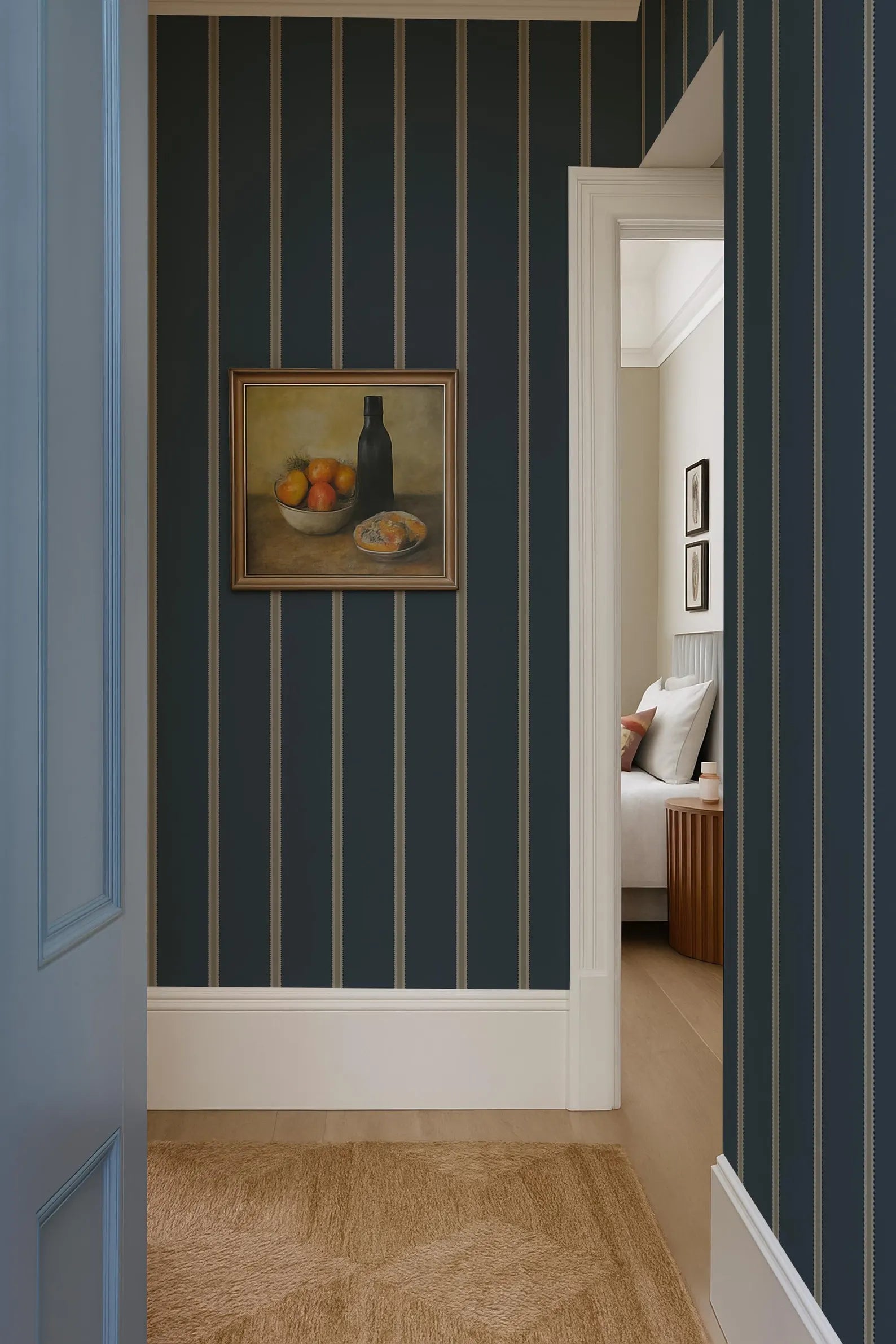 a hallway with a framed still life painting of fruit on the wall, a blue and white striped wallpaper, and a doorway leading to another room.