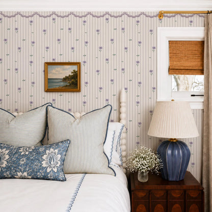 a cozy bedroom with a bed adorned with striped pillows and a blue and white floral bedspread. A wooden nightstand with a lamp and a vase of white flowers is situated next to the bed. The walls are decorated with a floral wallpaper, and a painting hangs above the bed.