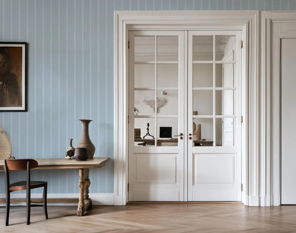 a room with a wooden table, a vase, and a chair. The room has a blue and white color scheme, with a large window and white doors.
