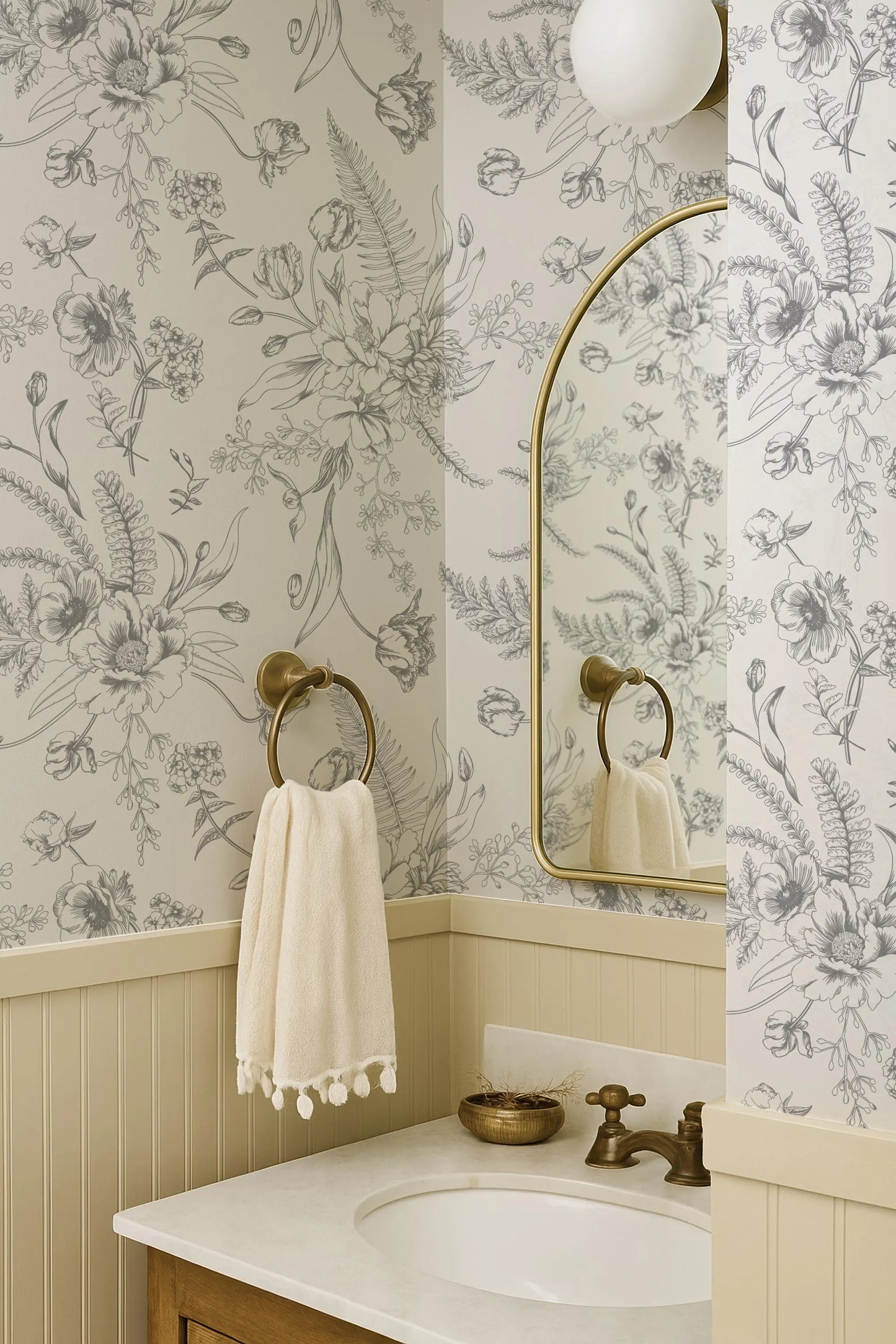 a bathroom with a white sink, a mirror, and a towel rack. The walls are decorated with a floral wallpaper, and there is a gold faucet on the sink.
