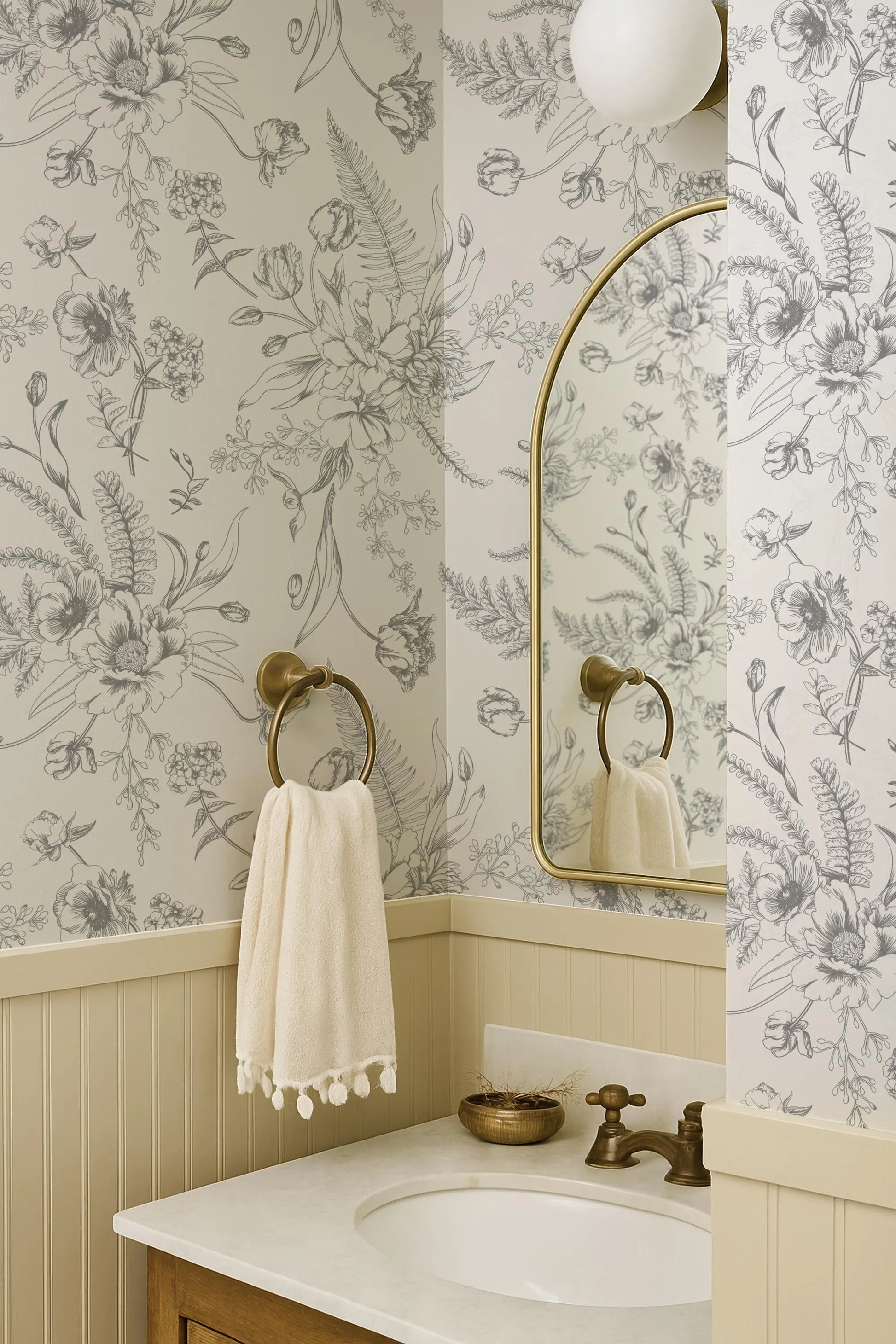a bathroom with a white sink, a mirror, and a towel rack. The walls are decorated with a floral wallpaper, and there is a gold faucet on the sink.