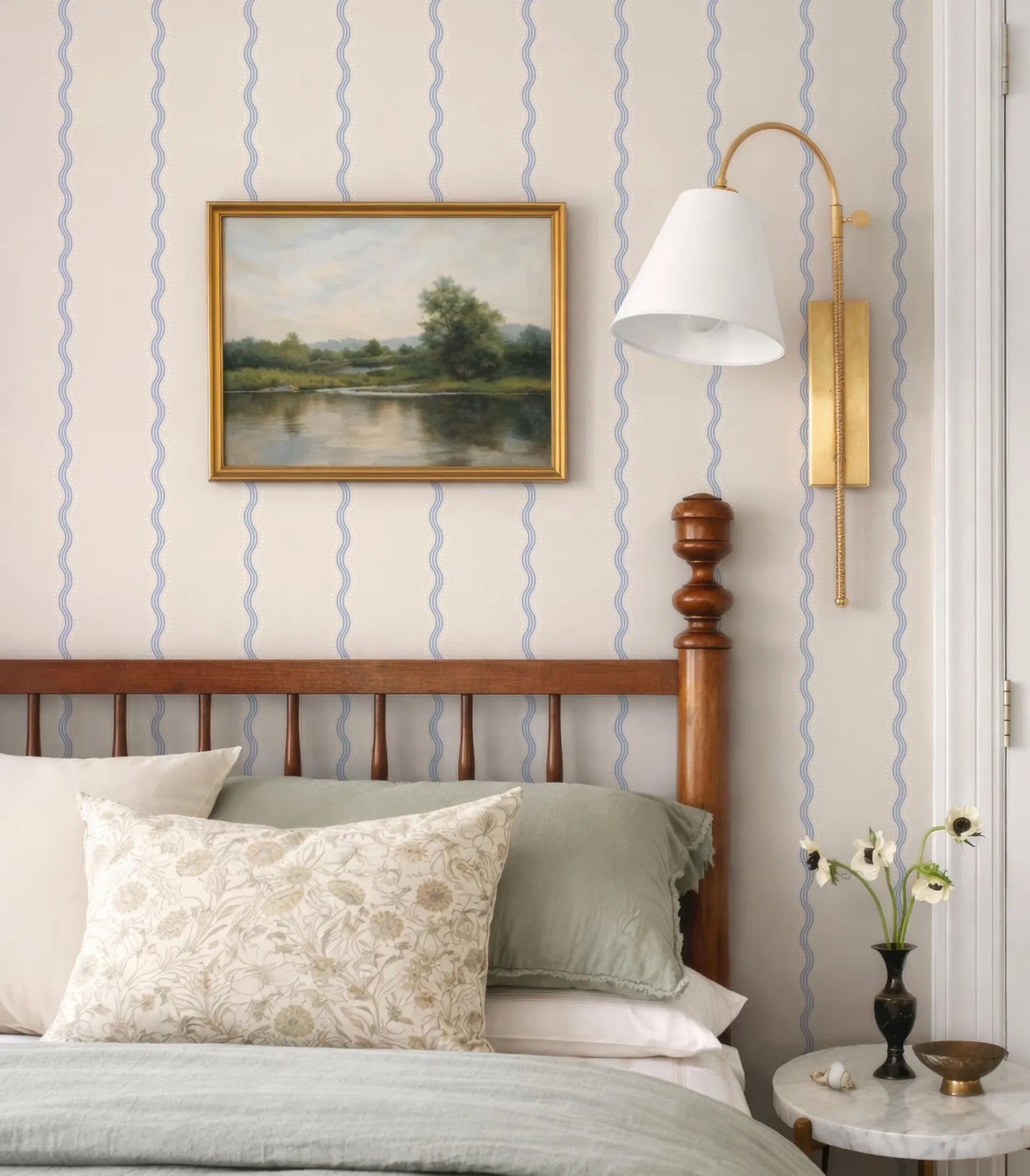a bedroom with a wooden headboard, a framed landscape painting on the wall, a lamp with a white shade, and a small table with a vase of flowers.