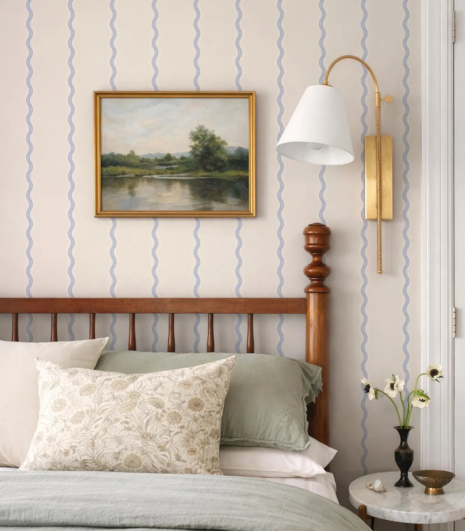 a bedroom with a wooden headboard, a framed landscape painting on the wall, a lamp with a white shade, and a small table with a vase of flowers.